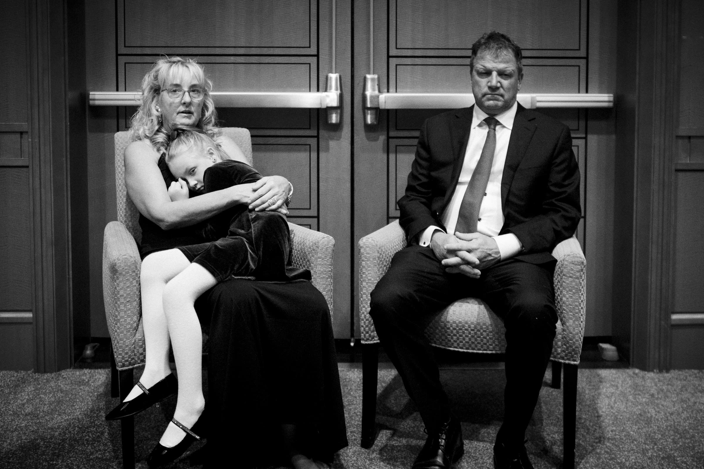 In a room with wooden doors, conveying a somber or reflective atmosphere, a woman cradles a sleeping child, while a man in a suit sits with his hands clasped and eyes closed. The scene unfolds like an intimate moment at an Umstead Hotel wedding.