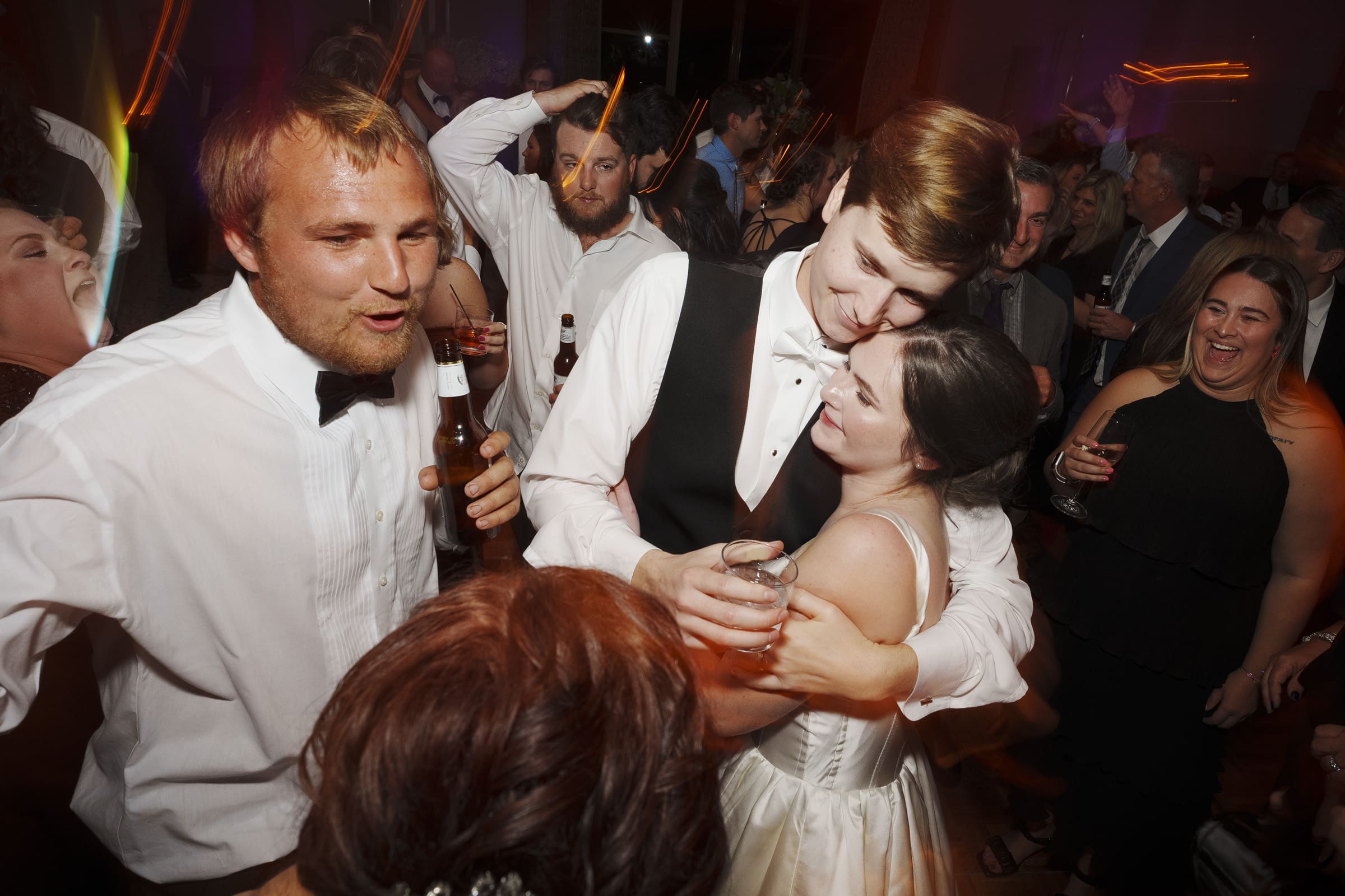At an Umstead Hotel wedding, a bride and groom dance closely in a lively crowd at the reception. The groom, in his vest, and the bride, in her gown, sway surrounded by guests who are smiling, holding drinks, and reveling in the joyous celebration.