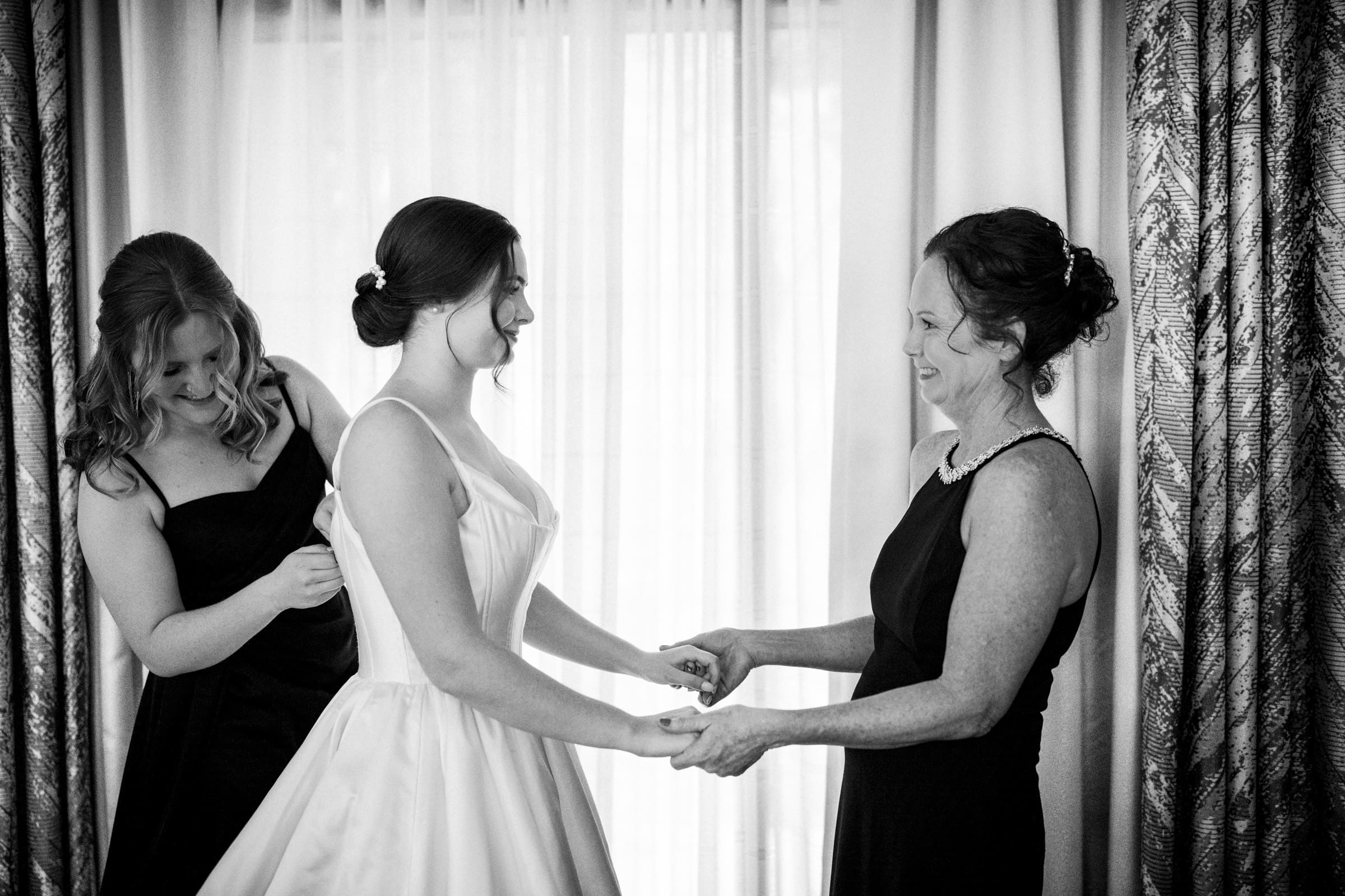 At an Umstead Hotel Wedding, a bride in a white dress stands before a curtain, holding hands with a woman in a dark dress. Another woman in a dark dress lovingly adjusts the gown from behind. All are smiling joyously.