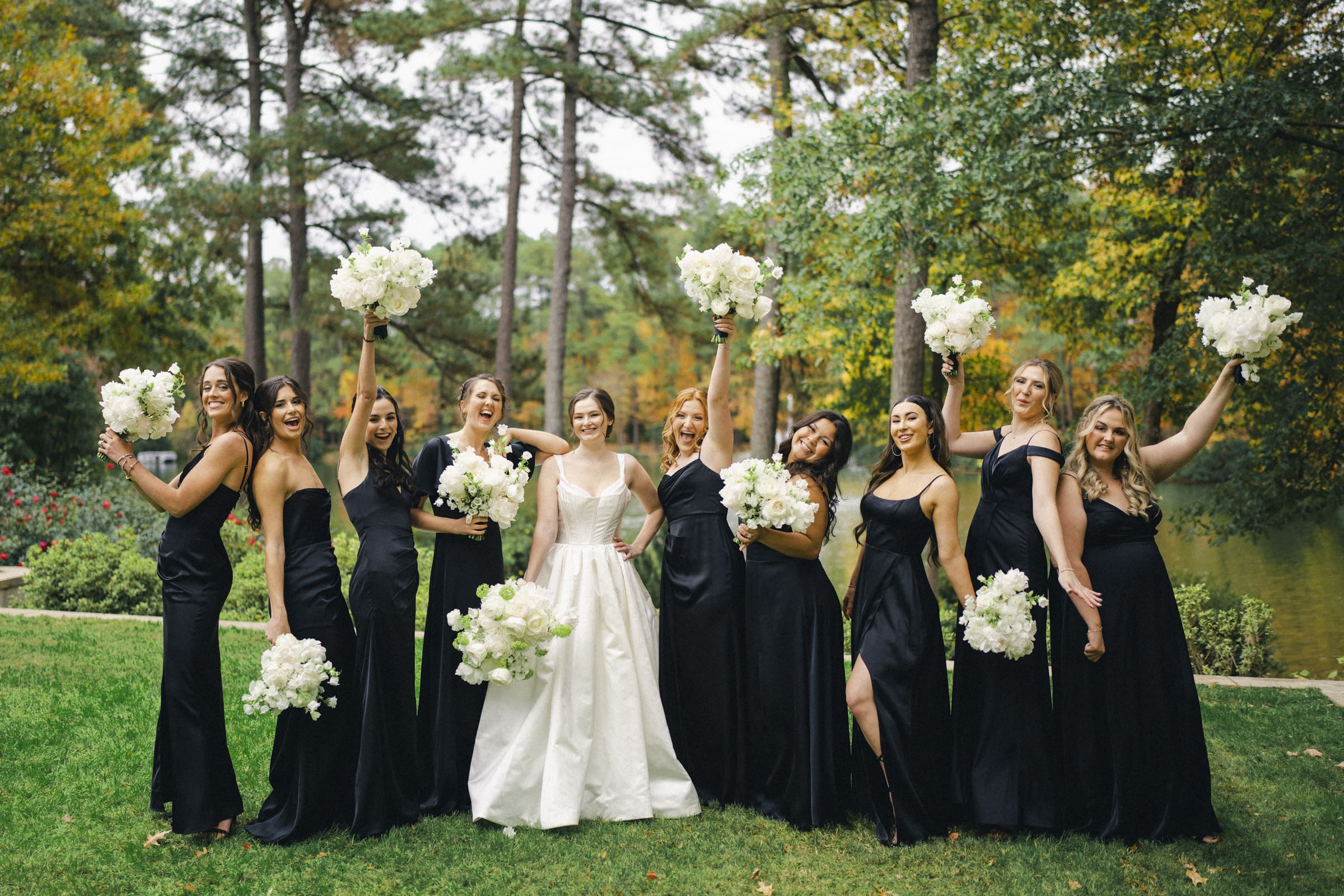 A bride in a white gown stands with bridesmaids in black dresses, all holding white flower bouquets. Celebrating an Umstead Hotel wedding, they pose outdoors on grass amidst tall trees, with some bridesmaids raising their bouquets joyfully.