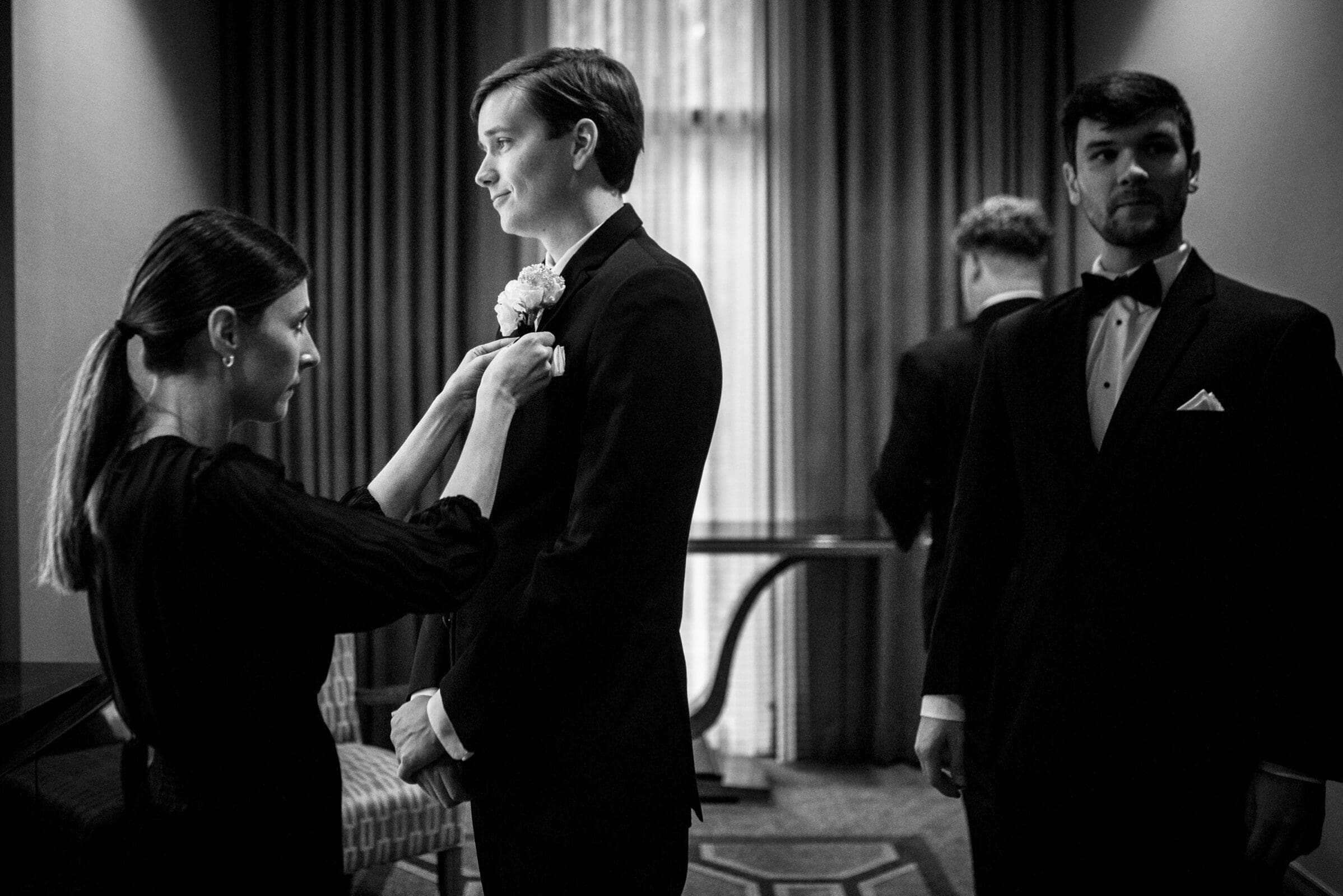 In a warmly lit room with curtains, a woman adjusts a boutonniere on a young man in a tuxedo, while another man in formal attire stands nearby. The scene exudes the elegance of an Umstead Hotel wedding, capturing the anticipation of the upcoming formal event.