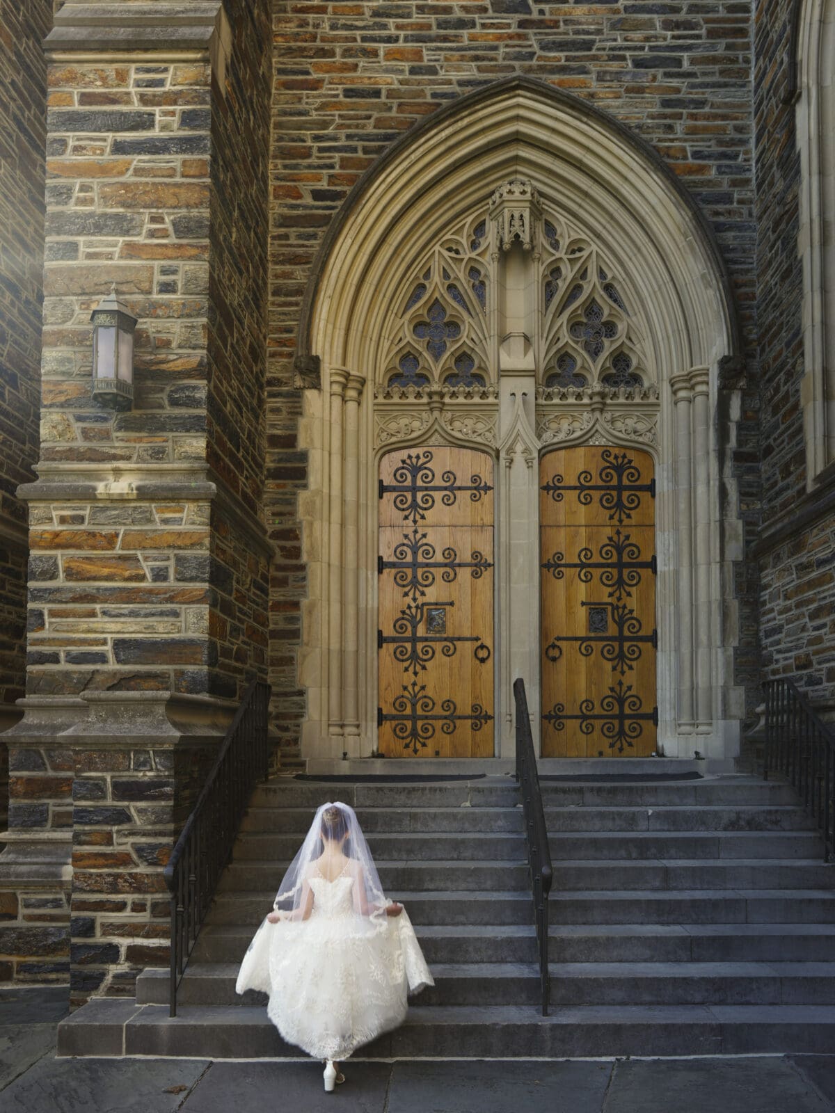 A bride in a white gown and veil stands on steps leading to an ornate, large wooden door of a stone building with arched detailing. Like a first holy communion portrait, she faces the door, her hands holding her dress, evoking a sense of anticipation and grandeur.