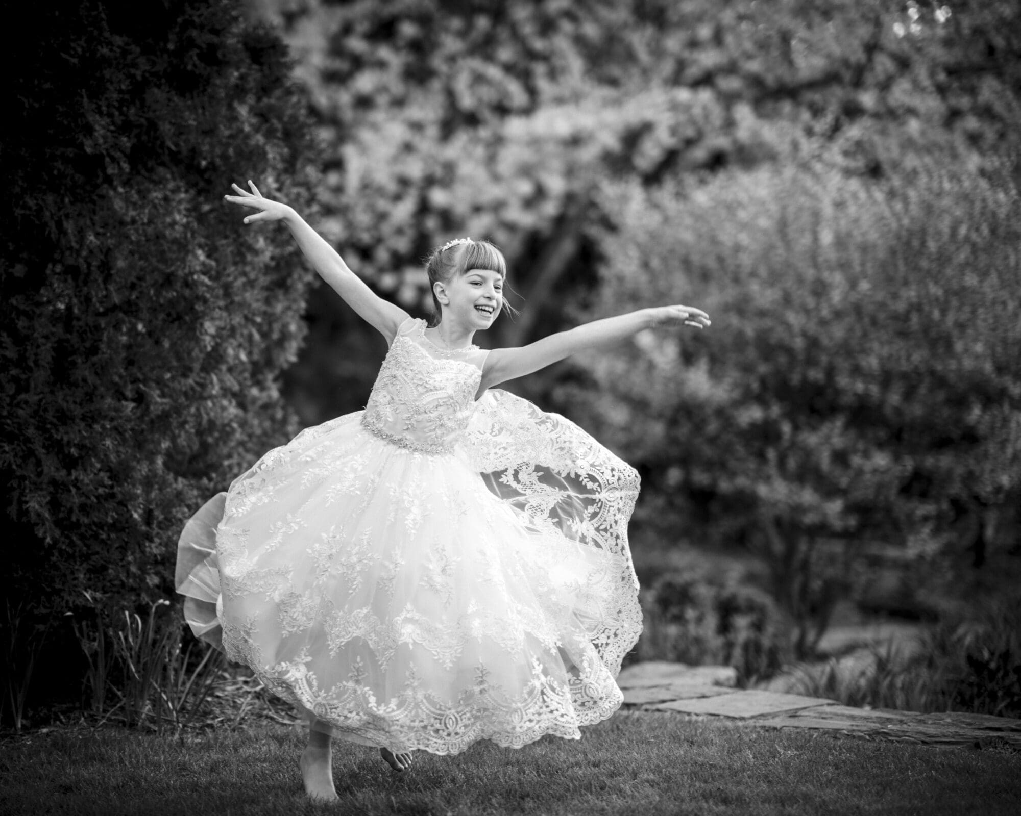 A young girl joyfully dances barefoot in a garden, her flowing lace dress reminiscent of first holy communion portrait photography. She has a bright smile, her arms gracefully extended. The black and white image captures the innocence framed by blurred lush greenery.