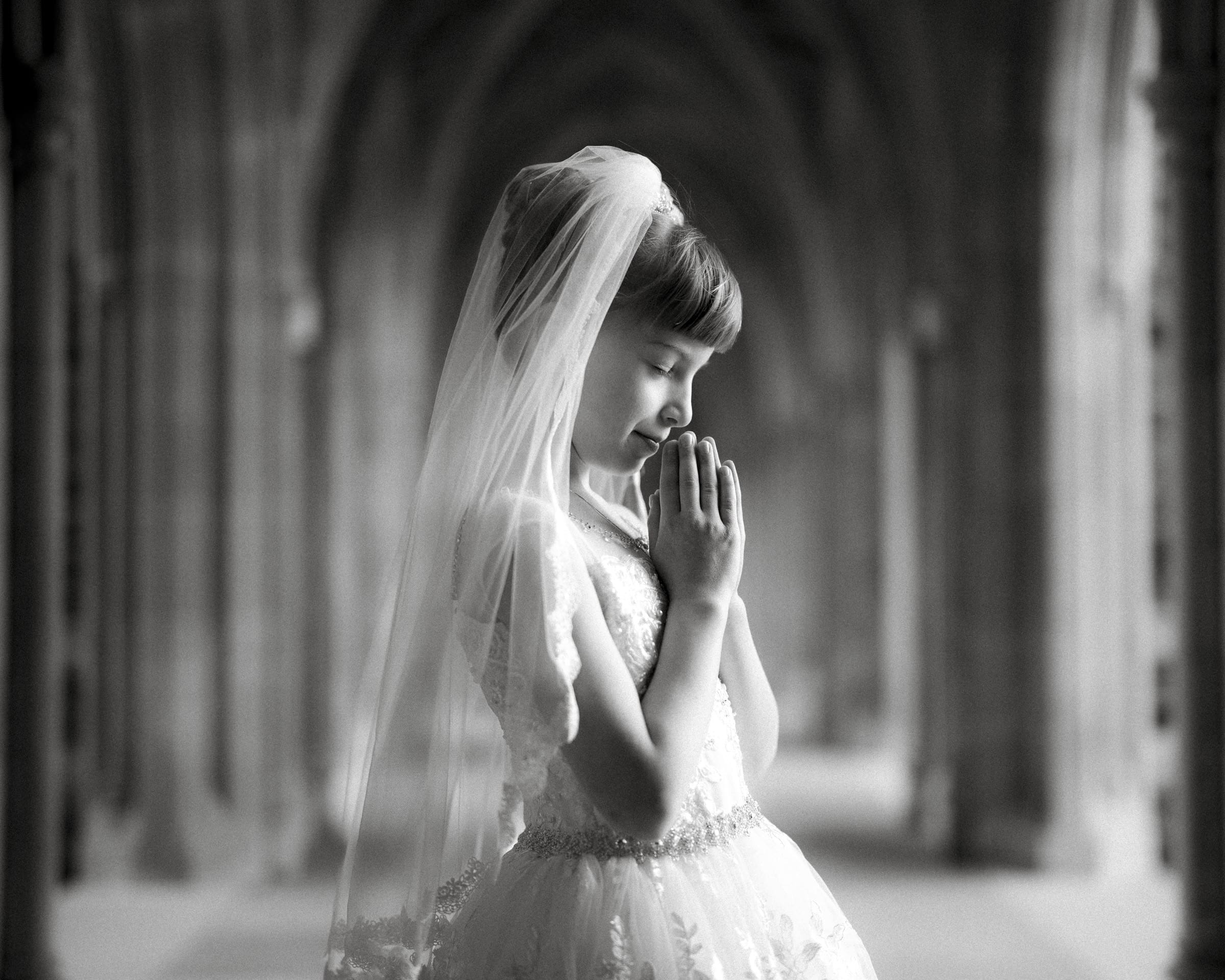 A young girl in a white dress and veil stands with her eyes closed and hands clasped in prayer, perfectly capturing the essence of first holy communion portrait photography. The serene and elegant setting within an ornate archway enhances the sense of a religious or ceremonial occasion.
