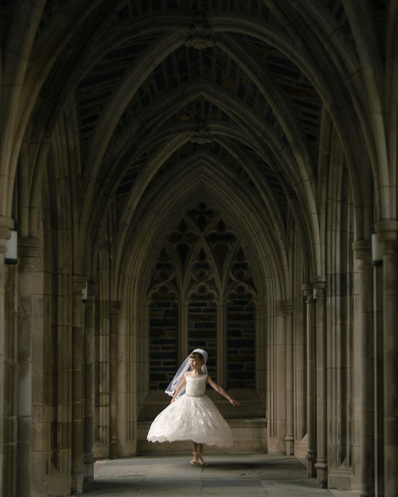 A bride in a white dress and veil dances gracefully in a dimly lit, gothic-style stone corridor with pointed arches and decorative windows, evoking the serene elegance of first holy communion portrait photography.
