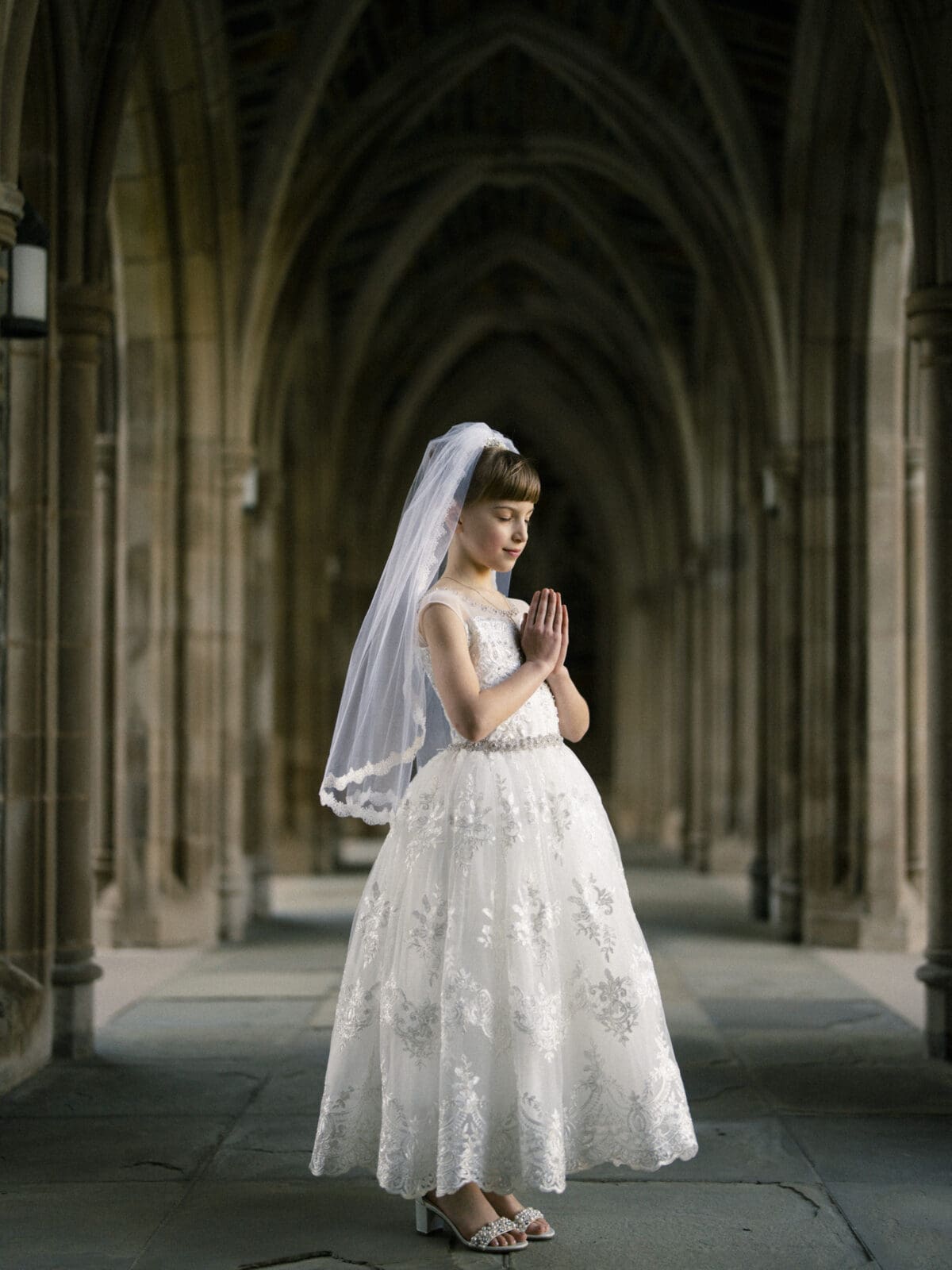 A young girl in a white lace communion dress and veil stands with hands clasped in prayer for her first holy communion portrait. She is framed by a stone archway with ornate columns, creating a serene and contemplative atmosphere.