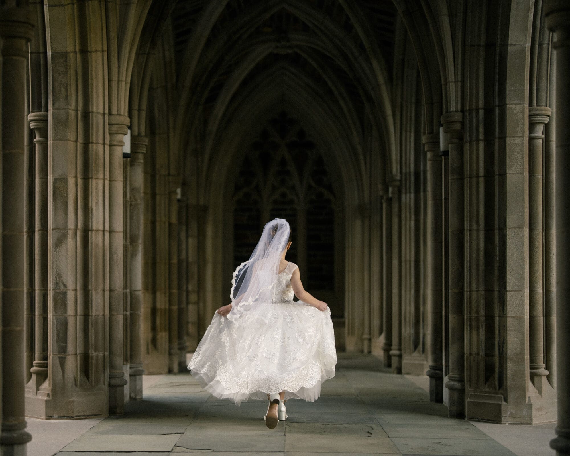 A bride in a white dress and veil walks down a stone hallway with arched ceilings, reminiscent of first holy communion portrait photography, holding up her gown as she moves forward. The setting is a dimly lit, gothic-style architectural passage.