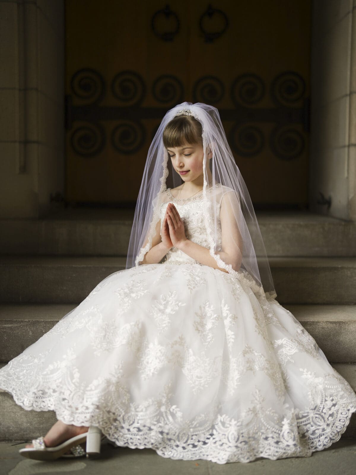 A young girl in a white lace dress and veil sits on stone steps, her hands clasped in prayer during her first holy communion. The ornate doors behind her enhance the portrait's solemn, serene atmosphere.