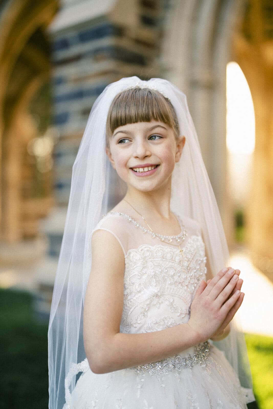 A young girl in a white dress and veil smiles while clasping her hands together in front of a stone building. Bathed in sunlight, the scene captures the essence of first holy communion portrait photography, with her joy radiating as she dresses for this special occasion.