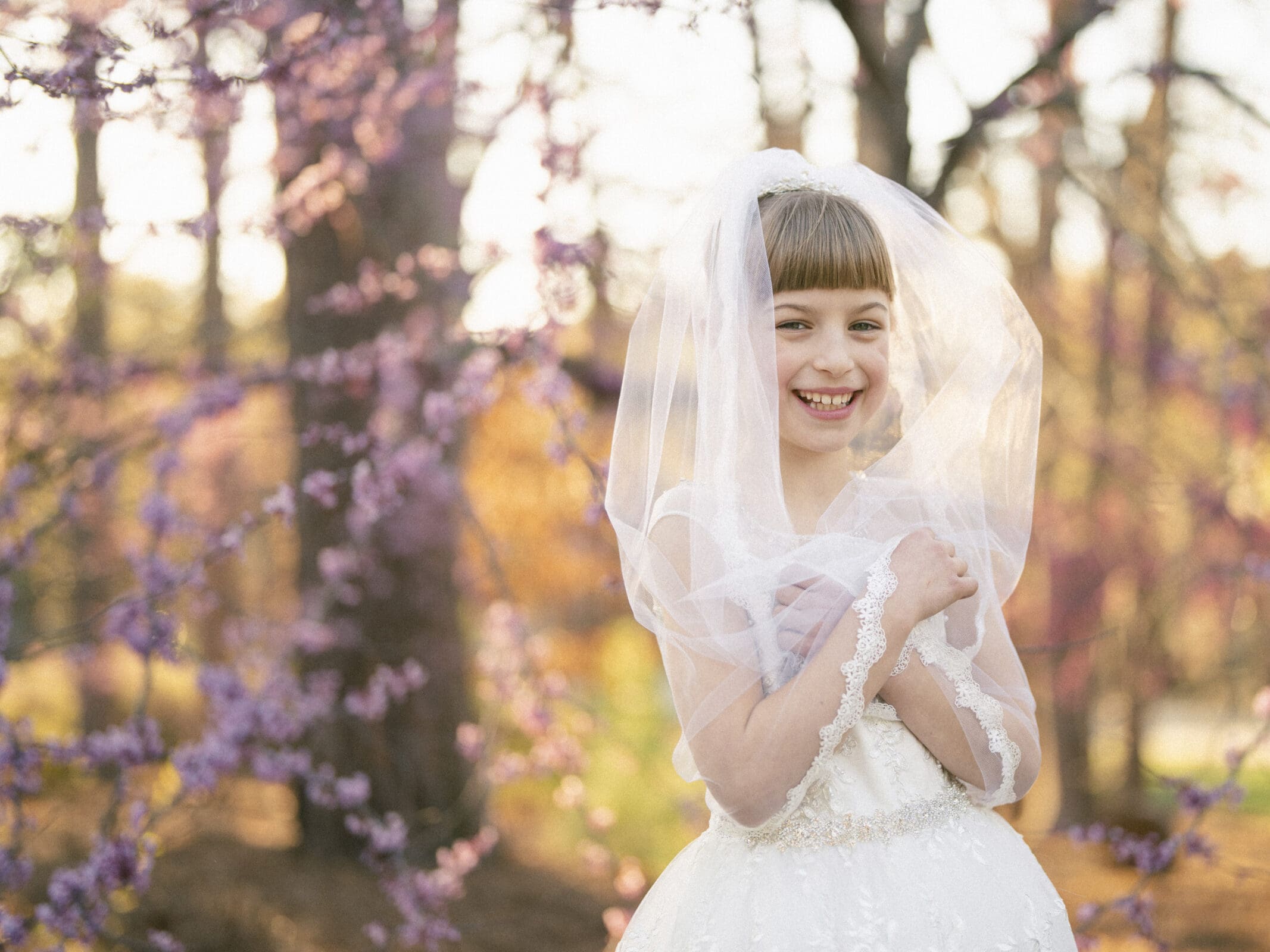 A young girl in a white dress and veil smiles joyfully, captured outdoors among blooming trees with purple flowers. The sunlight creates a warm, soft glow in this first holy communion portrait, epitomizing the essence of serene photography.