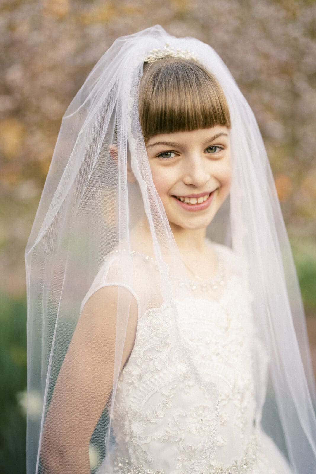 A young girl smiles while wearing a white lace dress and a translucent veil, embodying the essence of first holy communion portrait photography, set against a blurred natural background.