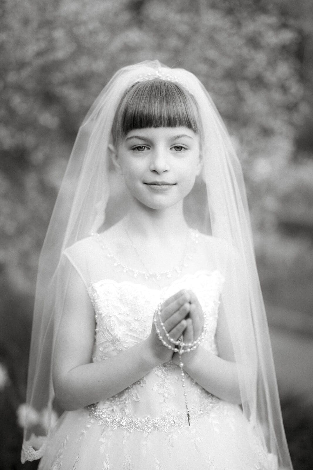 A young girl in a white lace dress and veil holds a rosary, capturing the essence of first holy communion portrait photography. She stands with her hands in prayer, the softly blurred background highlighting her serene expression. The image is in black and white.