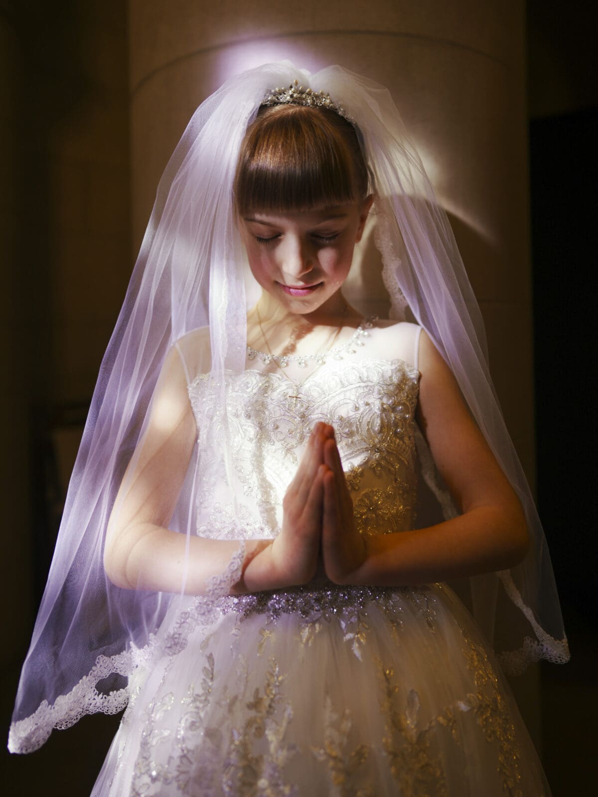 A young girl in an ornate white dress and veil clasps her hands in prayer, embodying a serene moment captured in first holy communion portrait photography. Her eyes are closed, with a delicate tiara gracing her head in the softly lit room.