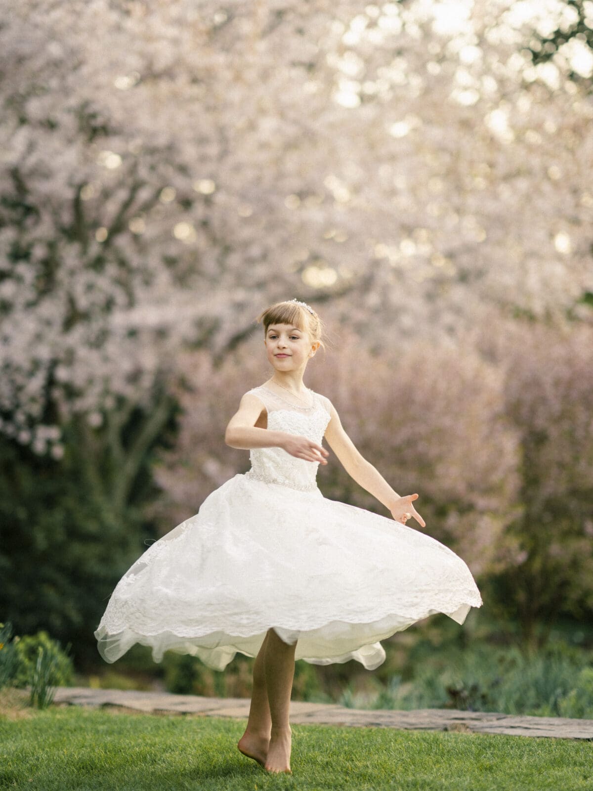 A girl in a white dress twirls on a grassy path surrounded by blooming trees, capturing the essence of first holy communion portrait photography. Her bare feet and the dress's movement emphasize a sense of freedom and joy, set against a backdrop filled with soft pink blossoms.