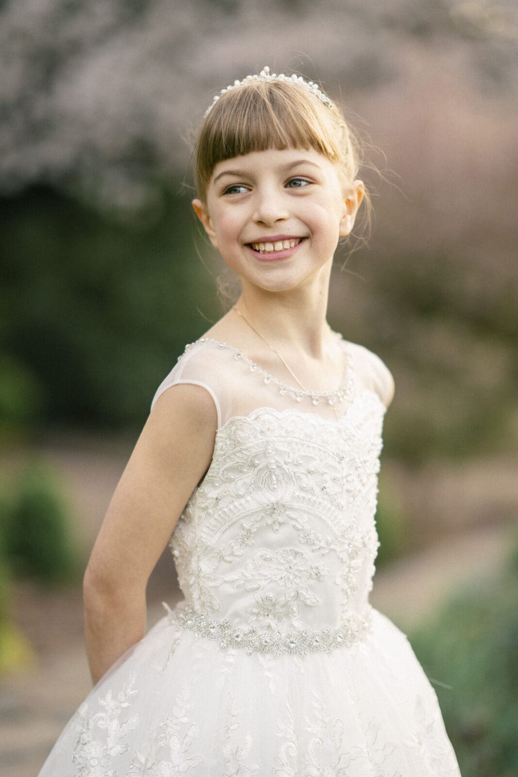 A young girl in a white, lace-embroidered dress with cap sleeves and a pearl headband smiles broadly while posing outdoors against a soft-focus background of trees, capturing the essence of first holy communion portrait photography.