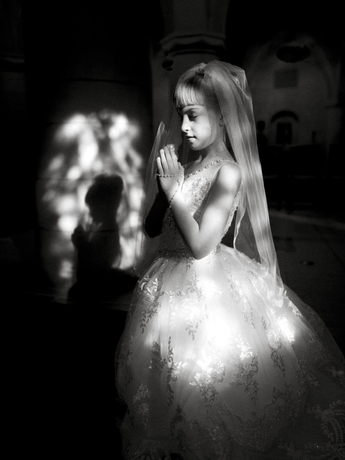 A young girl dressed in a white gown and veil stands with eyes closed and hands clasped in prayer, embodying the essence of a first holy communion portrait. Soft light filters through, creating patterns on her dress and casting her shadow on a nearby wall.