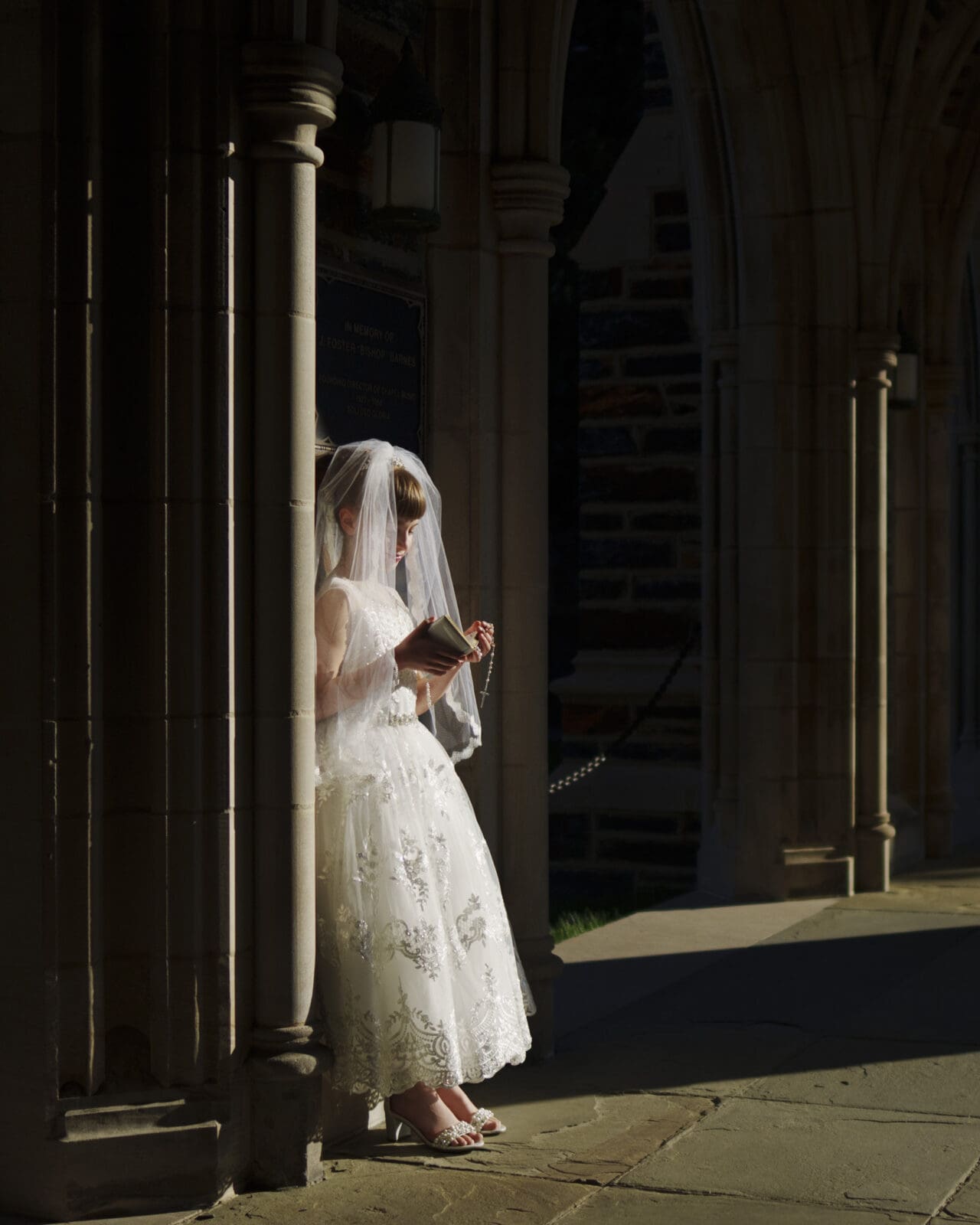 A bride in a white gown and veil stands under a stone archway, reading a book. Sunlight casts a gentle shadow, reminiscent of first holy communion portrait photography, highlighting the intricate lace details of her dress and the architectural elements around her.