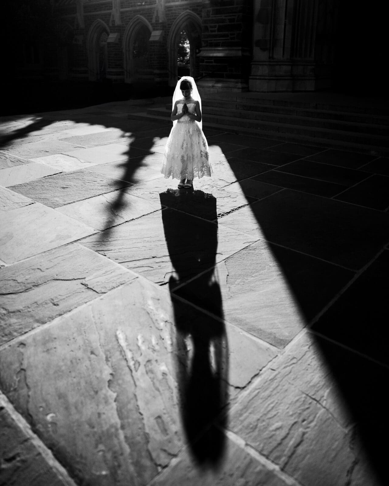 A young girl in a white dress and veil stands on a stone path, holding an object. Her shadow stretches dramatically in front of her, cast by strong sunlight, creating a striking contrast with the dark background—a beautiful moment of first holy communion portrait photography.