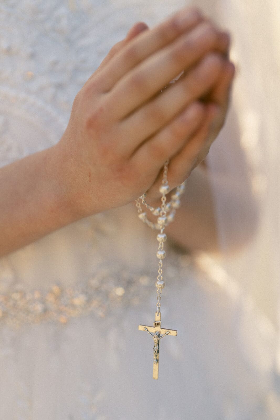 Close-up of a person in first holy communion portrait photography, holding a rosary with both hands in prayer. The rosary, featuring beads and a crucifix, complements the white garment adorned with intricate detailing.