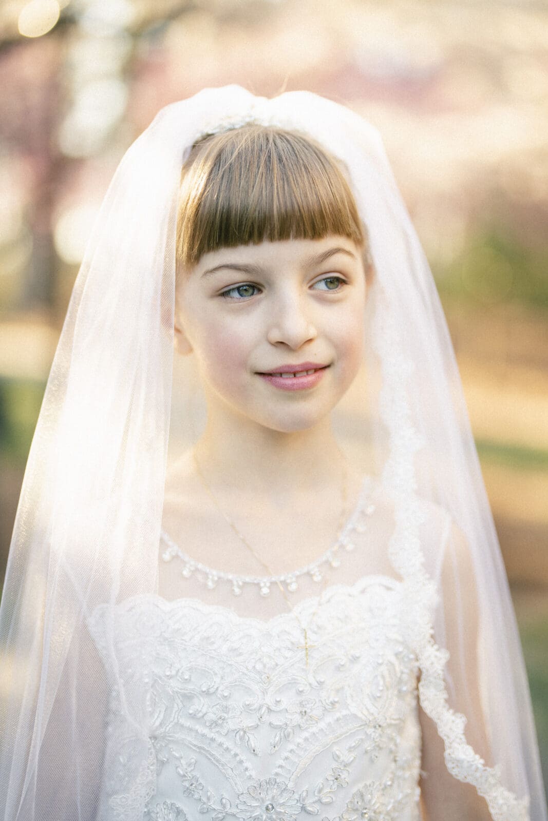 A young girl in a white dress and veil poses gracefully for a first holy communion portrait amidst a soft, blurred background of lush greenery and dappled sunlight.