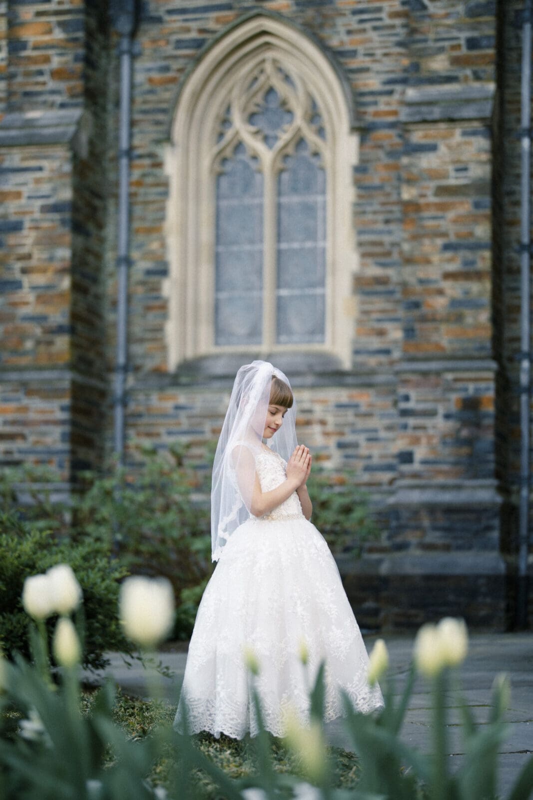 A young girl in a white dress and veil stands with hands clasped in prayer, capturing the essence of first holy communion portrait photography. She's near a stone church with arched windows, while white tulips blossom in the foreground.