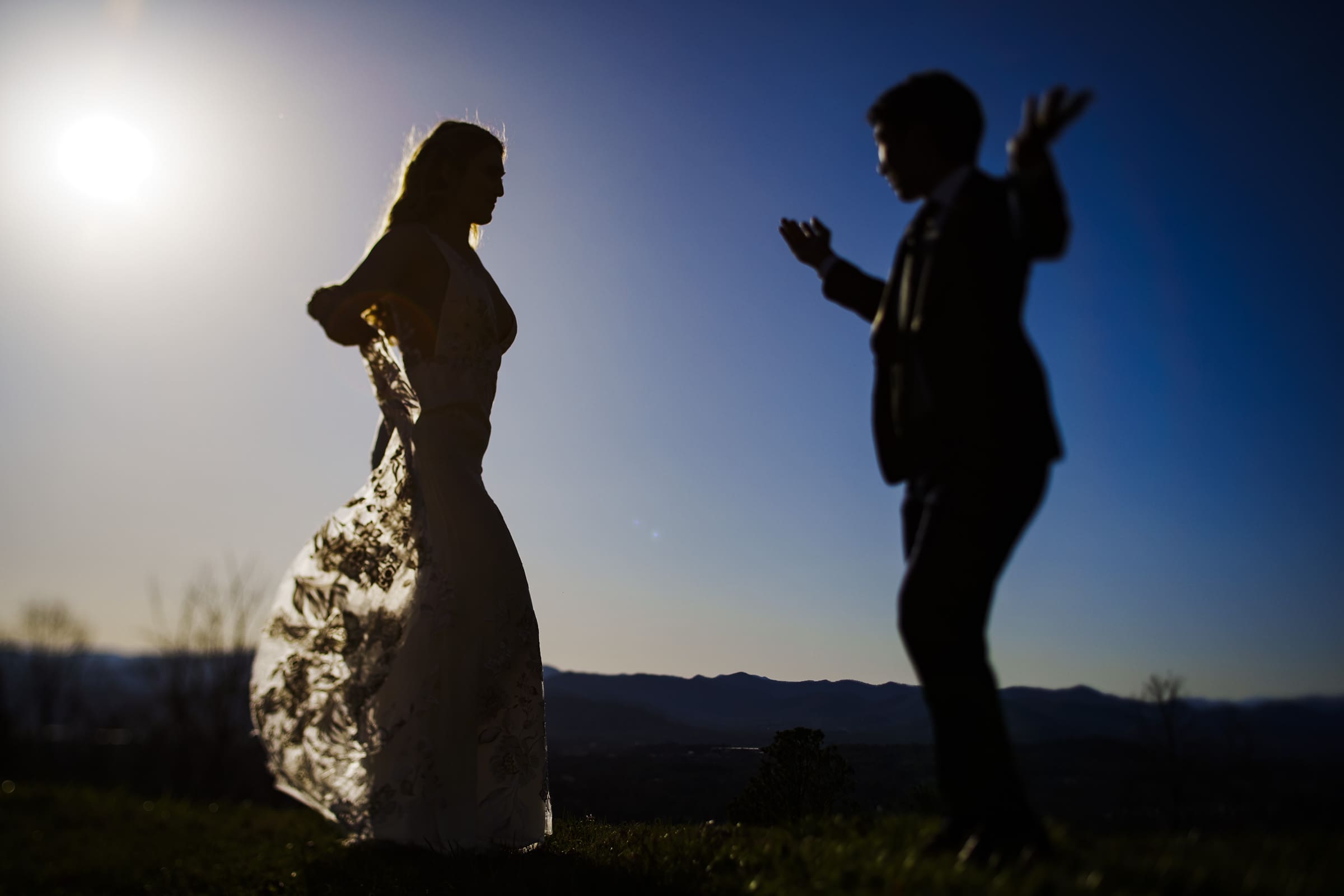 A silhouetted couple shares a tender moment outdoors, backlit by the sun. The woman in a flowing dress faces the man, who has his hands raised. Beyond them, the hills of Asheville set the scene for their intimate microwedding under a vast blue sky.