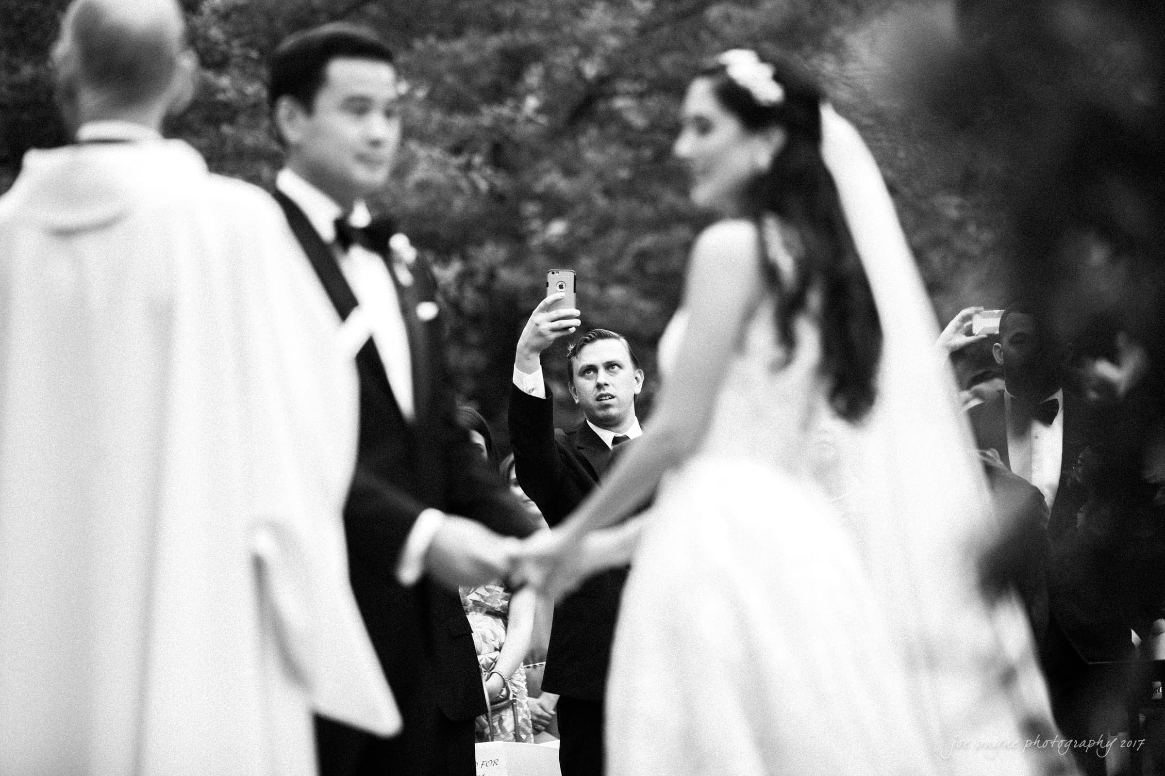 In an intimate, unplugged wedding ceremony, a couple exchanges vows outdoors. The groom and bride hold hands, facing each other tenderly. A man in the background captures the moment on his phone, dressed in a suit and looking intently. The timeless image is captured in black and white.