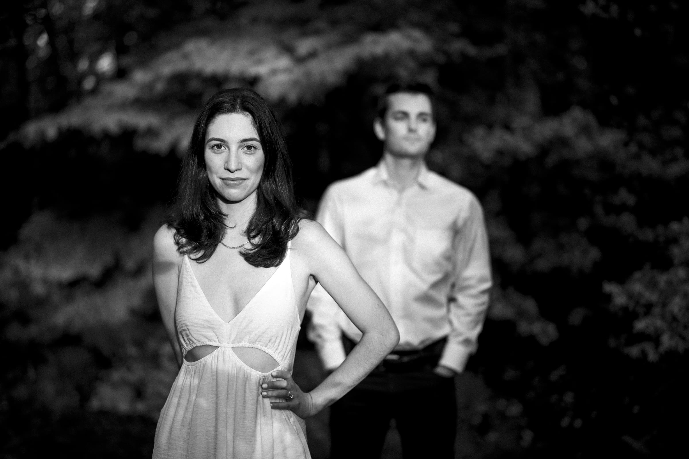 A black and white photo captures Gabby in a sleeveless dress standing in focus during the Duke Gardens engagement session, while Neil stands blurred in the background amidst a serene backdrop of trees.