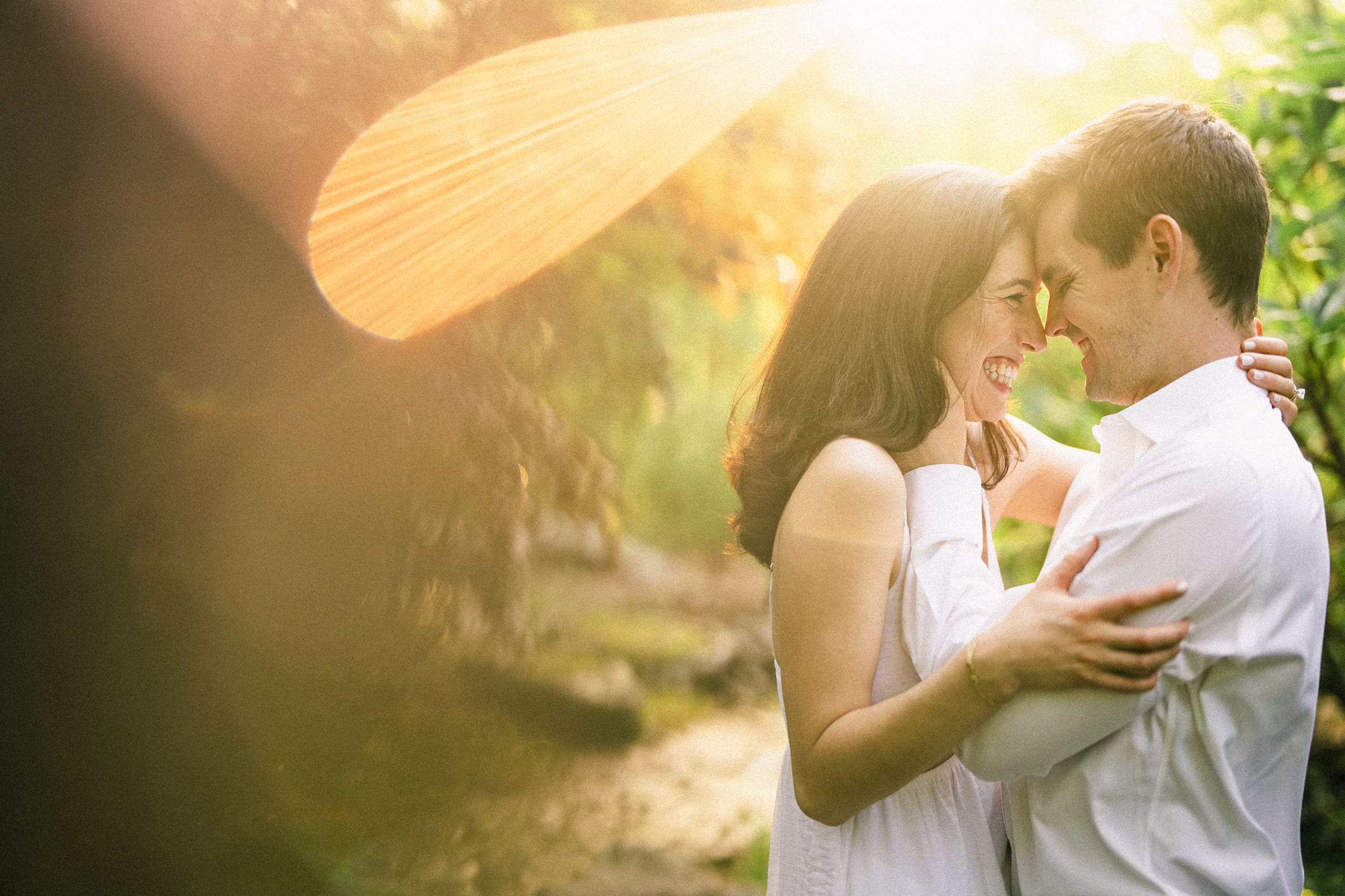 Gabby and Neil stand closely, smiling and embracing in a sunlit garden during their Duke Gardens engagement session. The sunlight filters through the trees, casting a warm, golden glow over their joyful expressions.