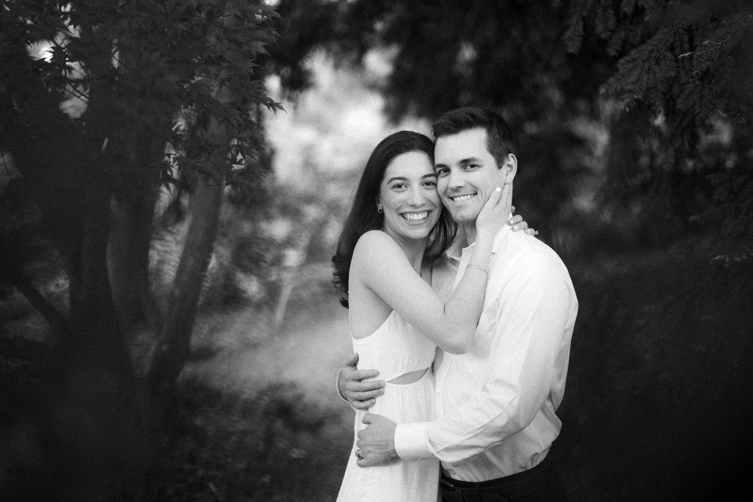 A black and white photo from a Duke Gardens engagement session captures Gabby embracing Neil. The couple's smiles light up the natural setting, as Neil dons a light-colored shirt. They are surrounded by blurred foliage, creating a soft, intimate atmosphere.