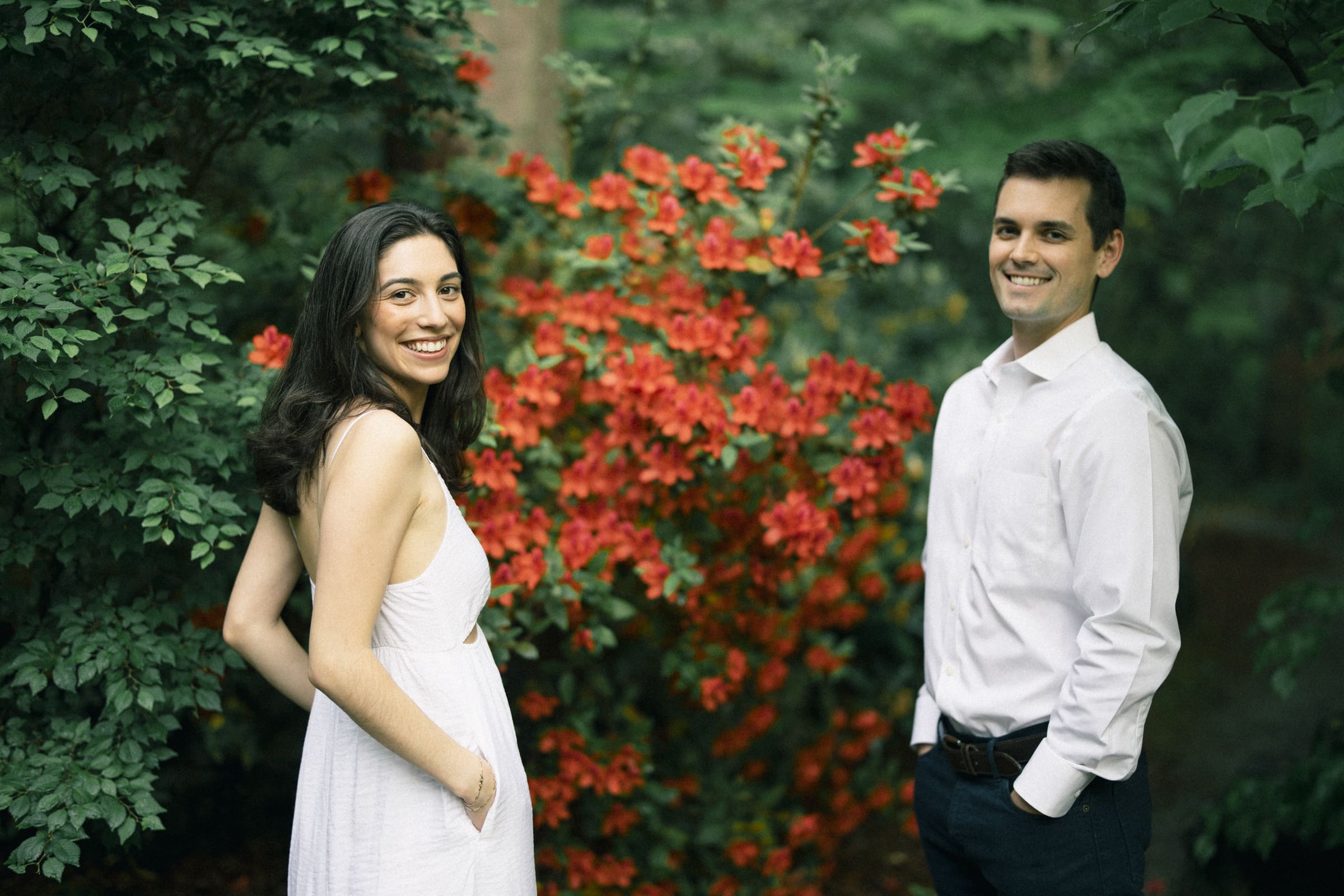 Gabby and Neil stand smiling in front of vibrant red flowers and lush greenery, their joy captured during a Duke Gardens engagement session. The setting is outdoors, suggesting a beautiful garden or park backdrop.