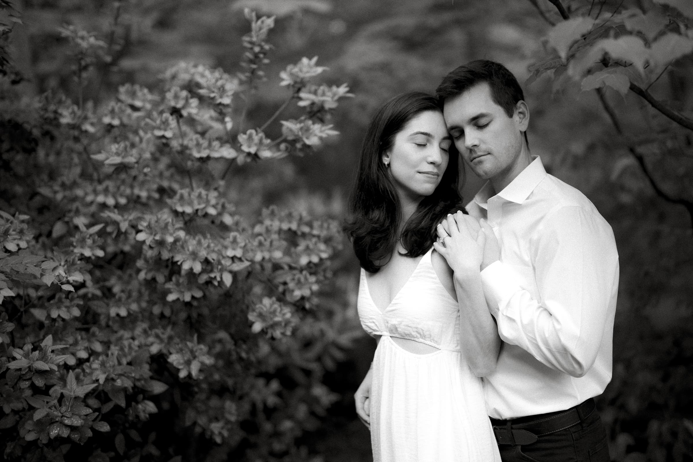 In a serene black and white image, Gabby and Neil stand closely in Duke Gardens, embracing tenderly. Dressed in white with relaxed expressions and eyes closed, they are surrounded by lush foliage that enhances the romantic atmosphere of their engagement session.