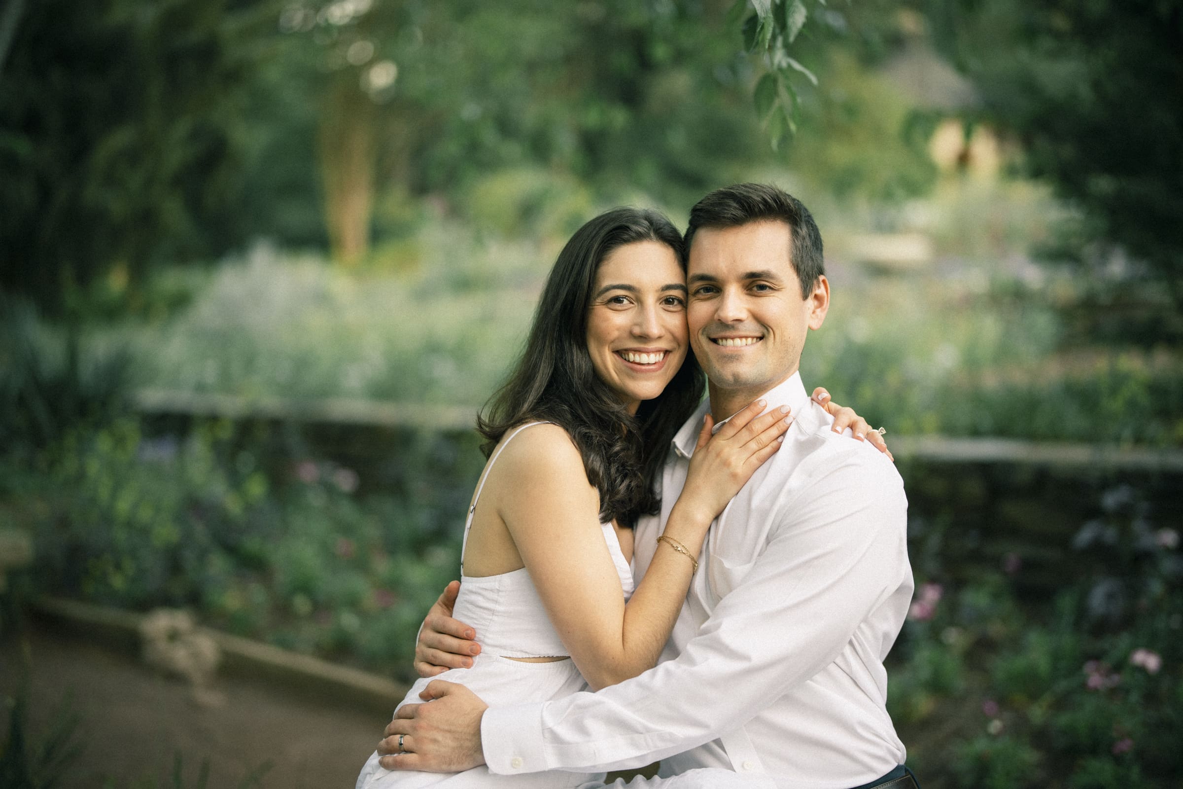 Gabby and Neil embrace and smile at the camera in a lush garden during their Duke Gardens engagement session. The woman wears a white dress, and the man a white shirt. The background is filled with greenery and soft-focus foliage, capturing their joy amidst nature's beauty.