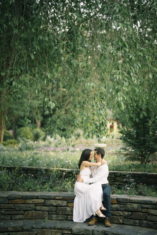 Gabby and Neil, dressed in white, sit closely together on a stone wall under hanging tree branches in a garden. They gaze into each other's eyes, surrounded by lush greenery and a tranquil atmosphere during their Duke Gardens engagement session.