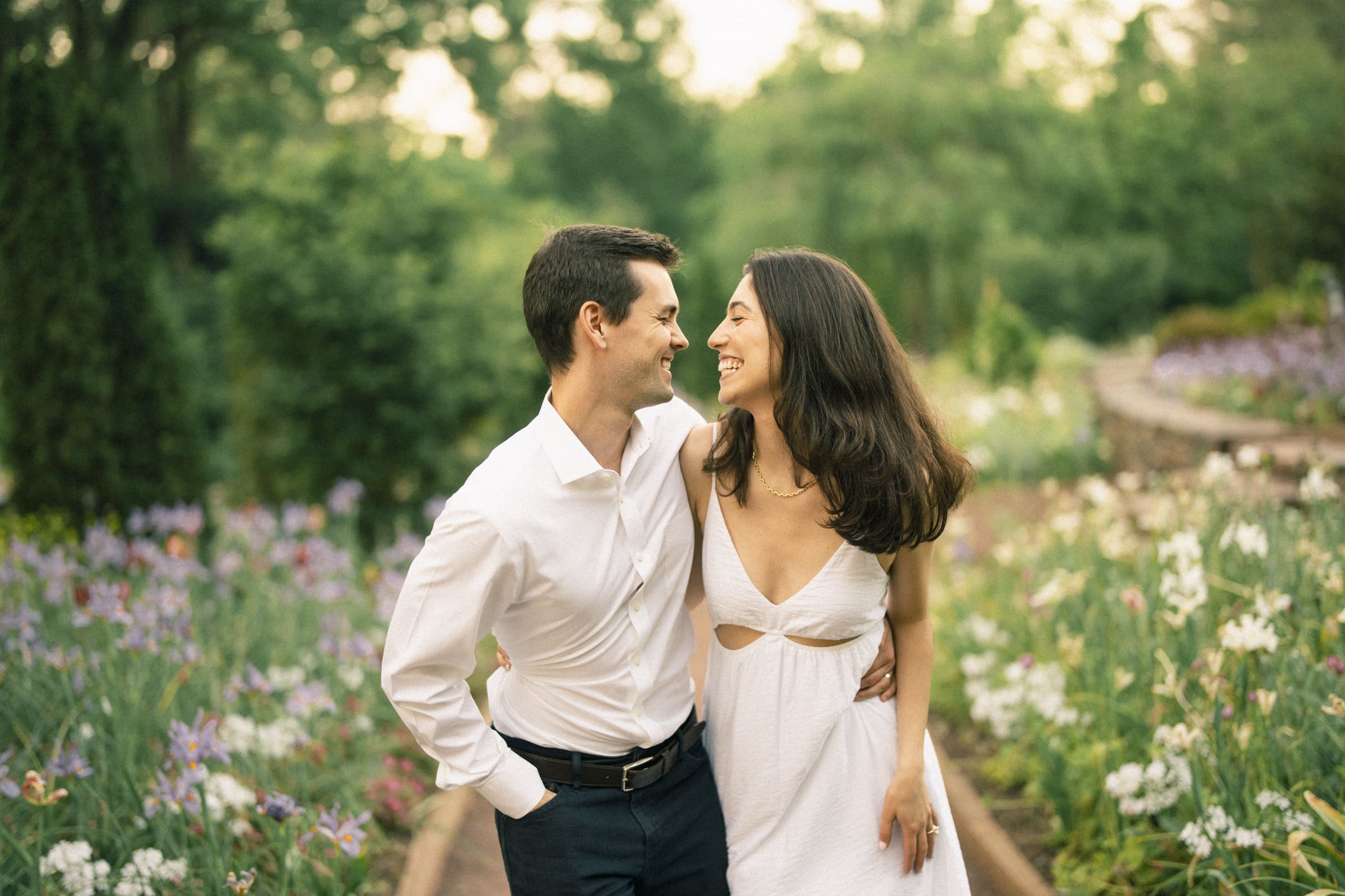 Gabby and Neil stand in Duke Gardens, enveloped by a vibrant array of colorful flowers. They smile warmly at each other; she in a white dress, he in a white shirt and dark pants. The greenery softly blurs into the background of their enchanting engagement session.