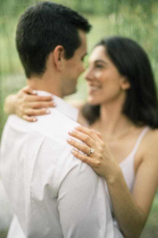 A couple embracing outdoors, Gabby and Neil smile softly at each other. The woman, with long hair in a white dress, shows off her engagement ring as she wraps her arms around his neck. The blurred greenery of Duke Gardens creates a beautiful backdrop for their engagement session.