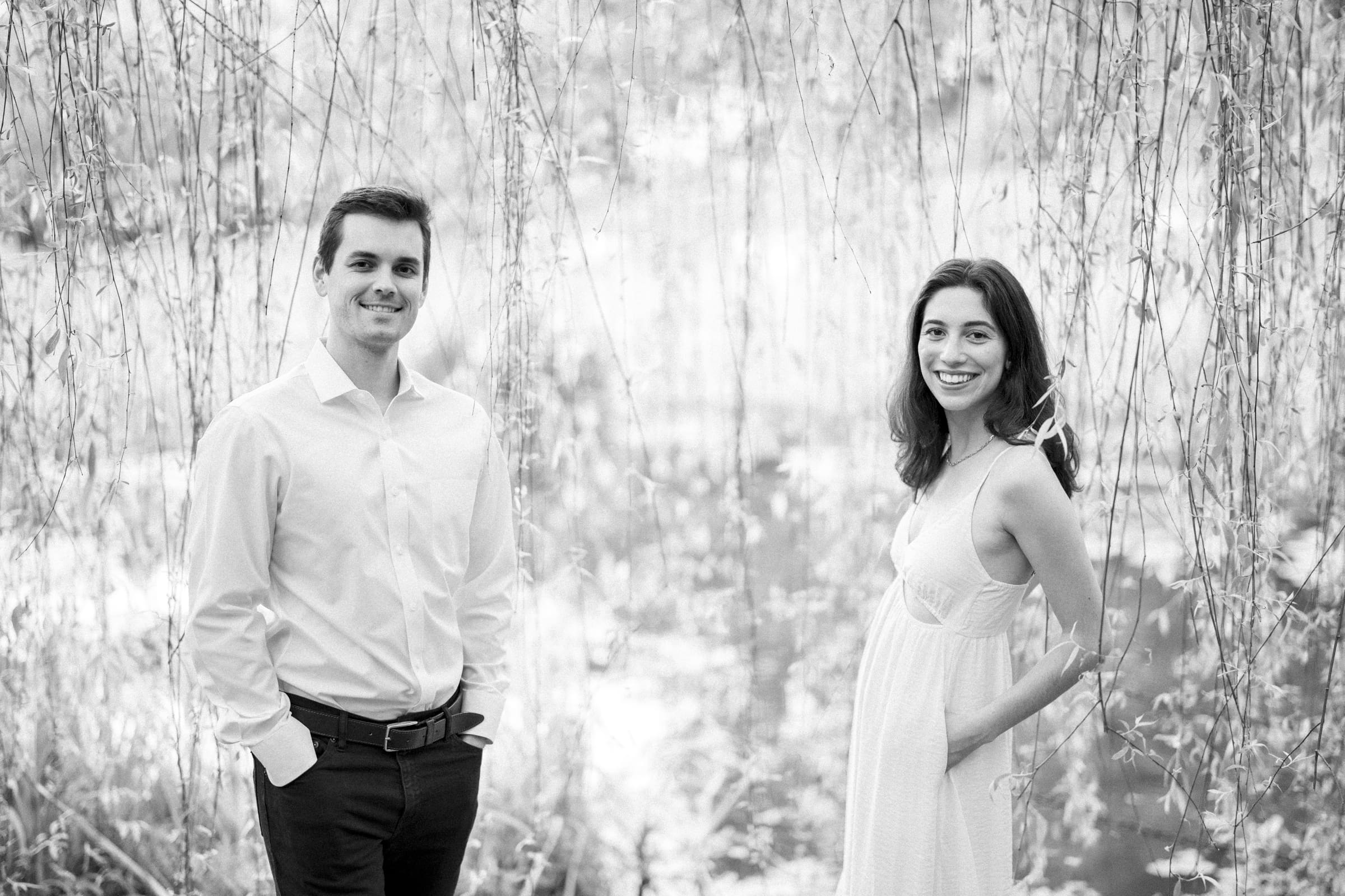 A black and white photo captures Gabby and Neil during their Duke Gardens engagement session. Among hanging branches, Neil stands on the left in a light shirt and dark pants with hands in pockets, while Gabby wears a light dress, smiling warmly at the camera.