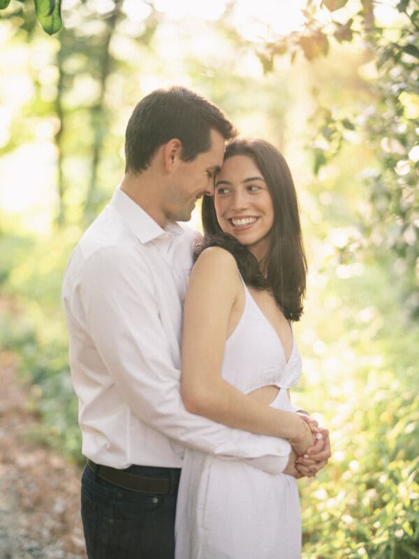 In a sunlit, leafy setting of Duke Gardens, Gabby and Neil share a moment of joy. She smiles in her white dress as he embraces her from behind. The blurred background beautifully highlights their joyful expressions during this enchanting engagement session.