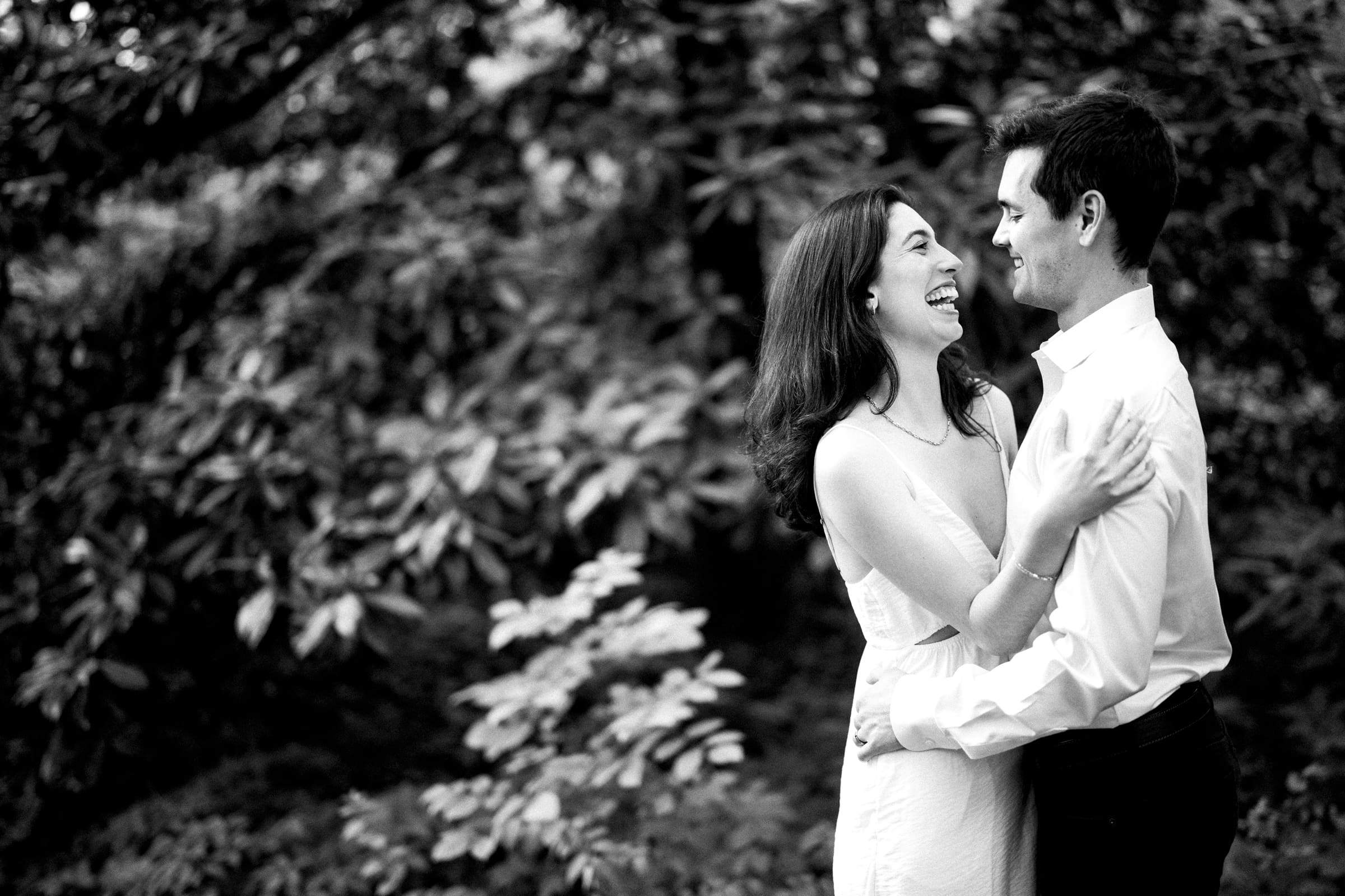 A black and white image captures Gabby and Neil joyfully embracing in Duke Gardens. The woman is in a light dress, the man in a white shirt. Both smile at each other, surrounded by lush foliage, perfectly illustrating their engagement session's tender moment.