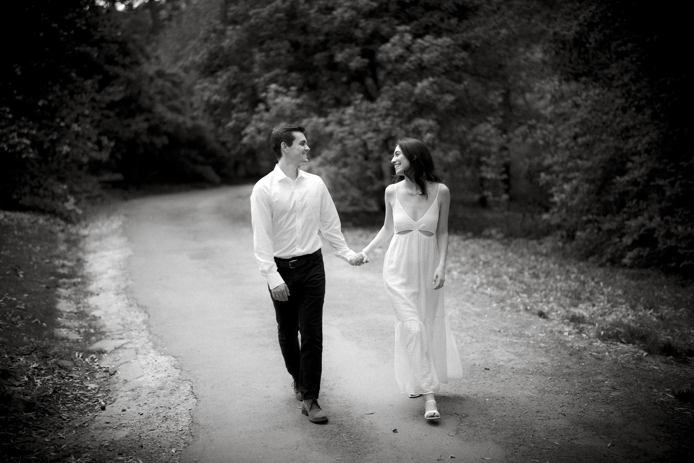 A black and white photo captures Gabby and Neil walking hand in hand along a tree-lined path during their Duke Gardens engagement session. The man wears a white shirt and dark pants; the woman, in a flowing white dress, looks at him. They appear happy and relaxed in the serene outdoor setting.