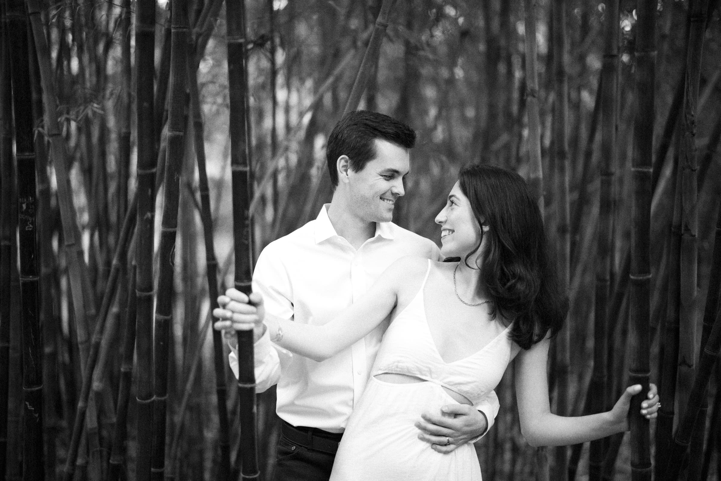 Gabby and Neil share a tender moment amidst tall bamboo stalks during their Duke Gardens engagement session. The man, wearing a white shirt, holds onto the bamboo as the woman in a light dress beamingly looks at him. The serene setting enhances their joyful expressions.