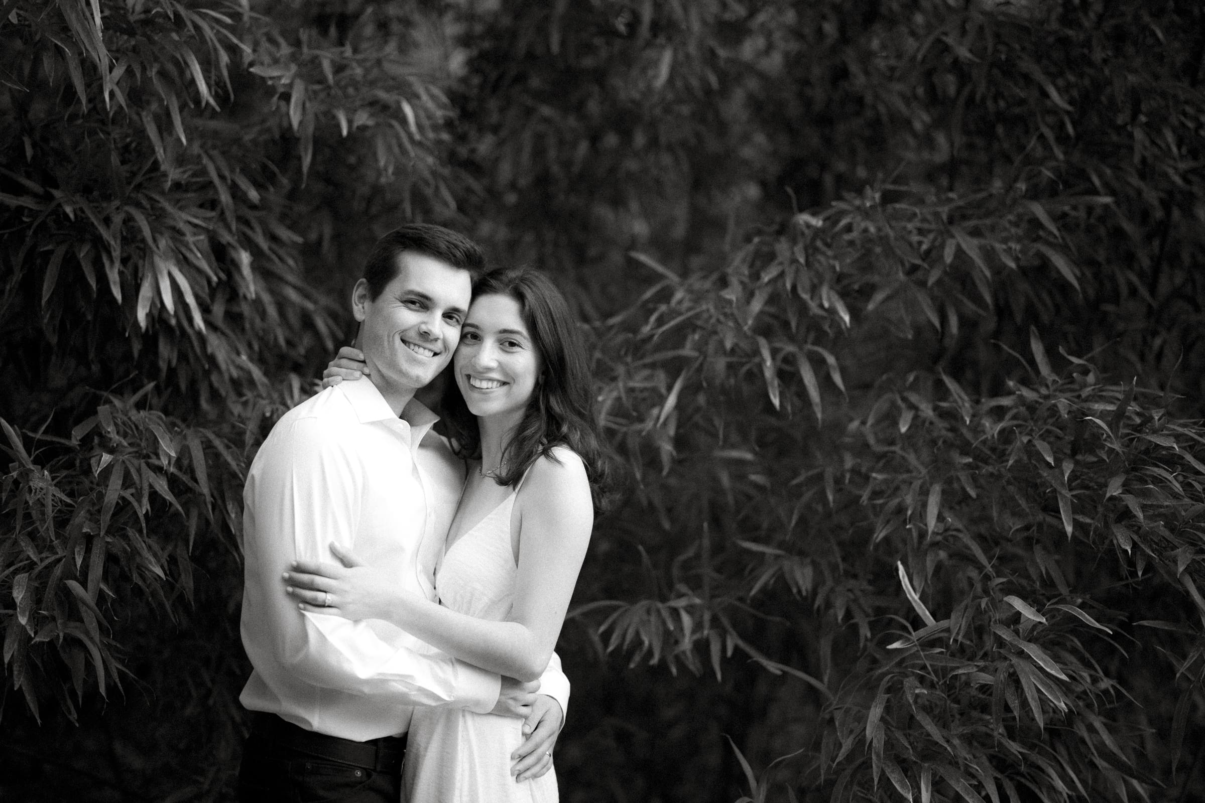 A black and white photo of Gabby and Neil, a smiling couple embracing amidst the lush foliage during their Duke Gardens engagement session. The man is wearing a light shirt, while the woman is in a sleeveless dress.