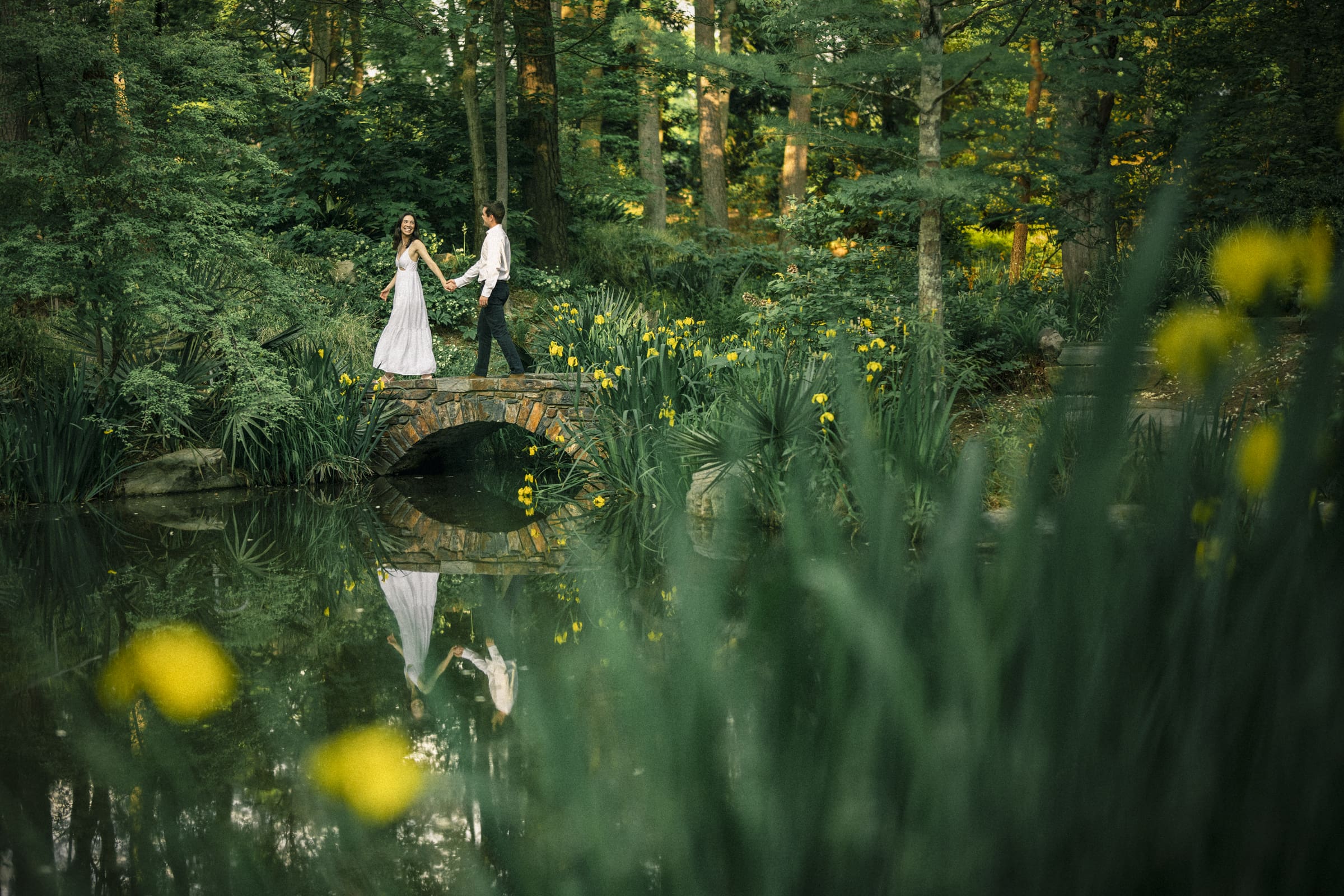 Gabby and Neil stroll hand in hand over a small stone bridge at Duke Gardens, she in a white dress and he in a white shirt and black pants. Surrounded by lush greenery and yellow flowers, their reflections ripple gently in the serene water, capturing the essence of their engagement session.