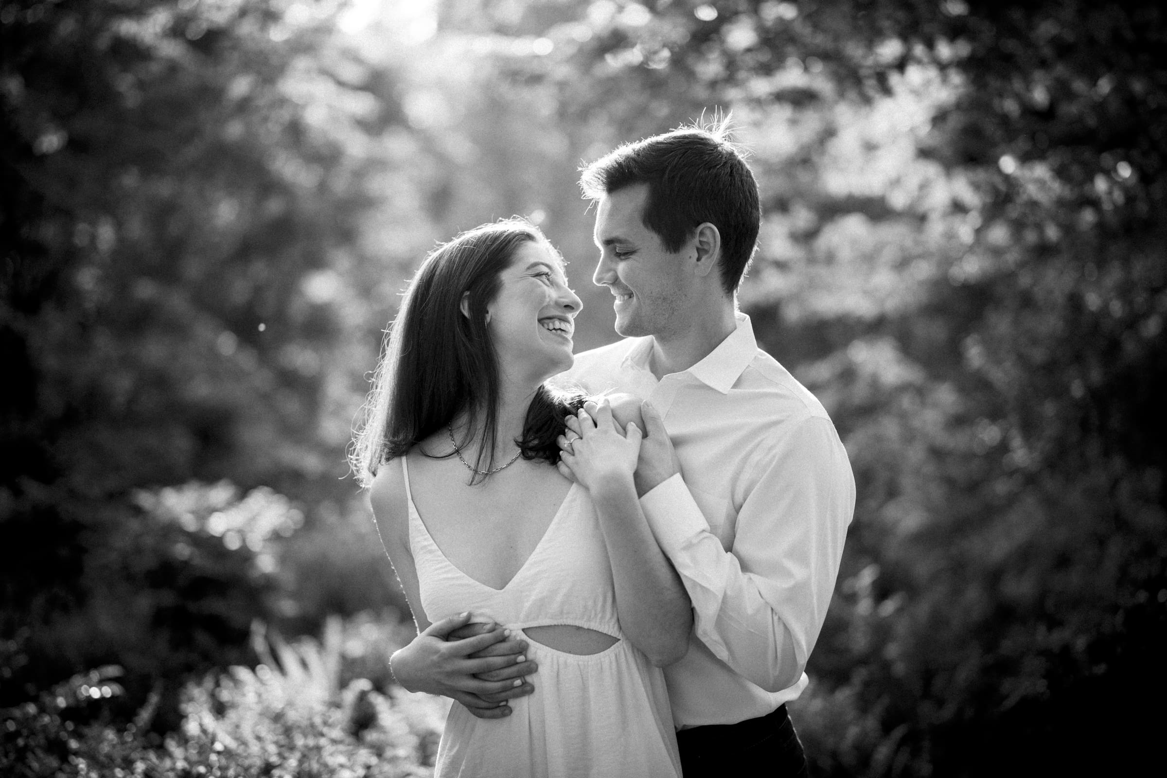 Gabby and Neil smile lovingly at each other in a sunlit forest during their Duke Gardens engagement session. The woman in a sleeveless dress gently holds the man's shoulder, while he wears a white shirt. Sunlight filters through the trees, creating a peaceful, romantic atmosphere.