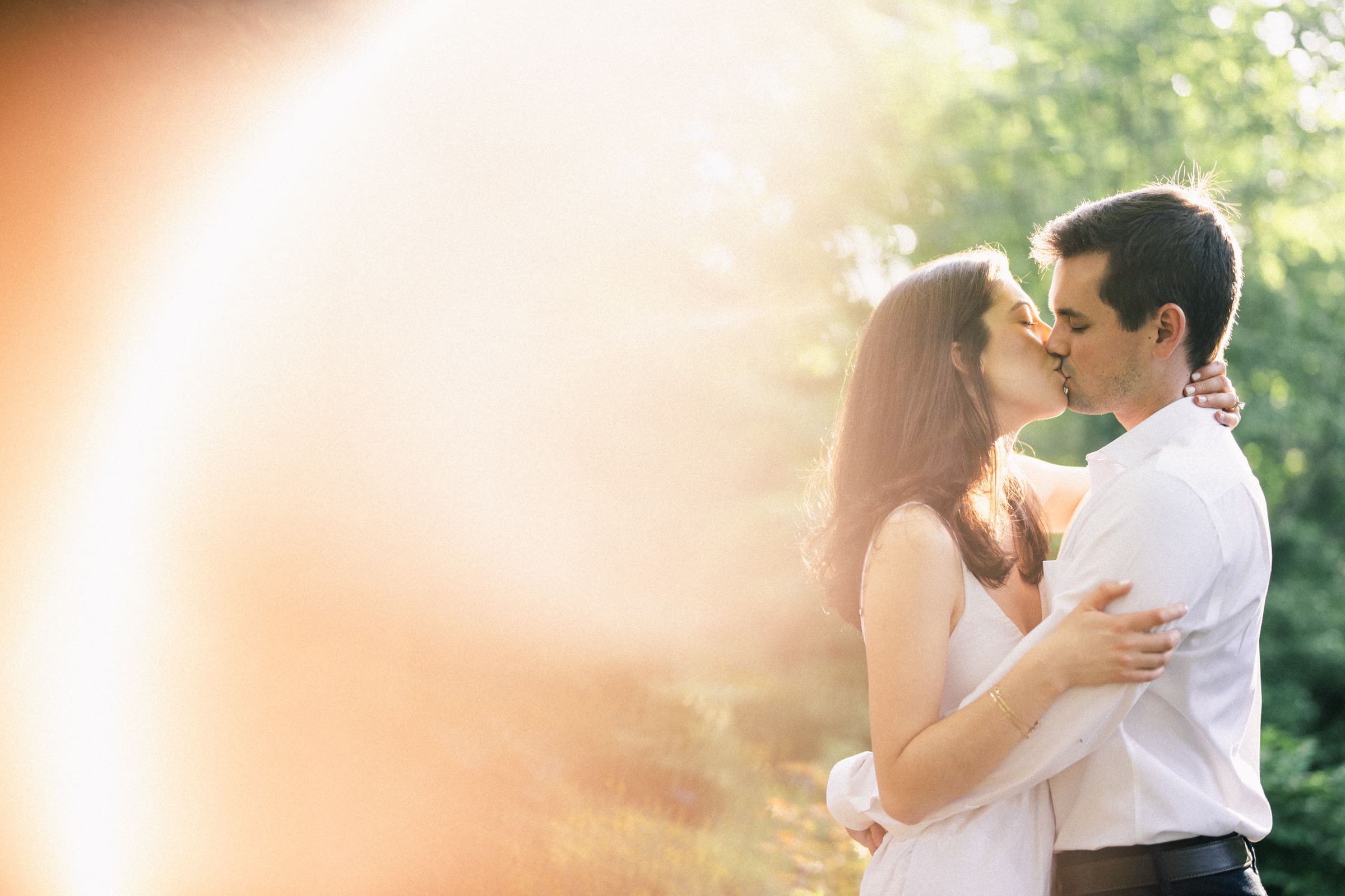 A couple kisses in a sunlit garden, surrounded by soft, blurred light. The woman wears a white dress, and the man wears a white shirt, embracing each other lovingly amid green foliage—capturing the essence of Gabby & Neil's Duke Gardens engagement session.
