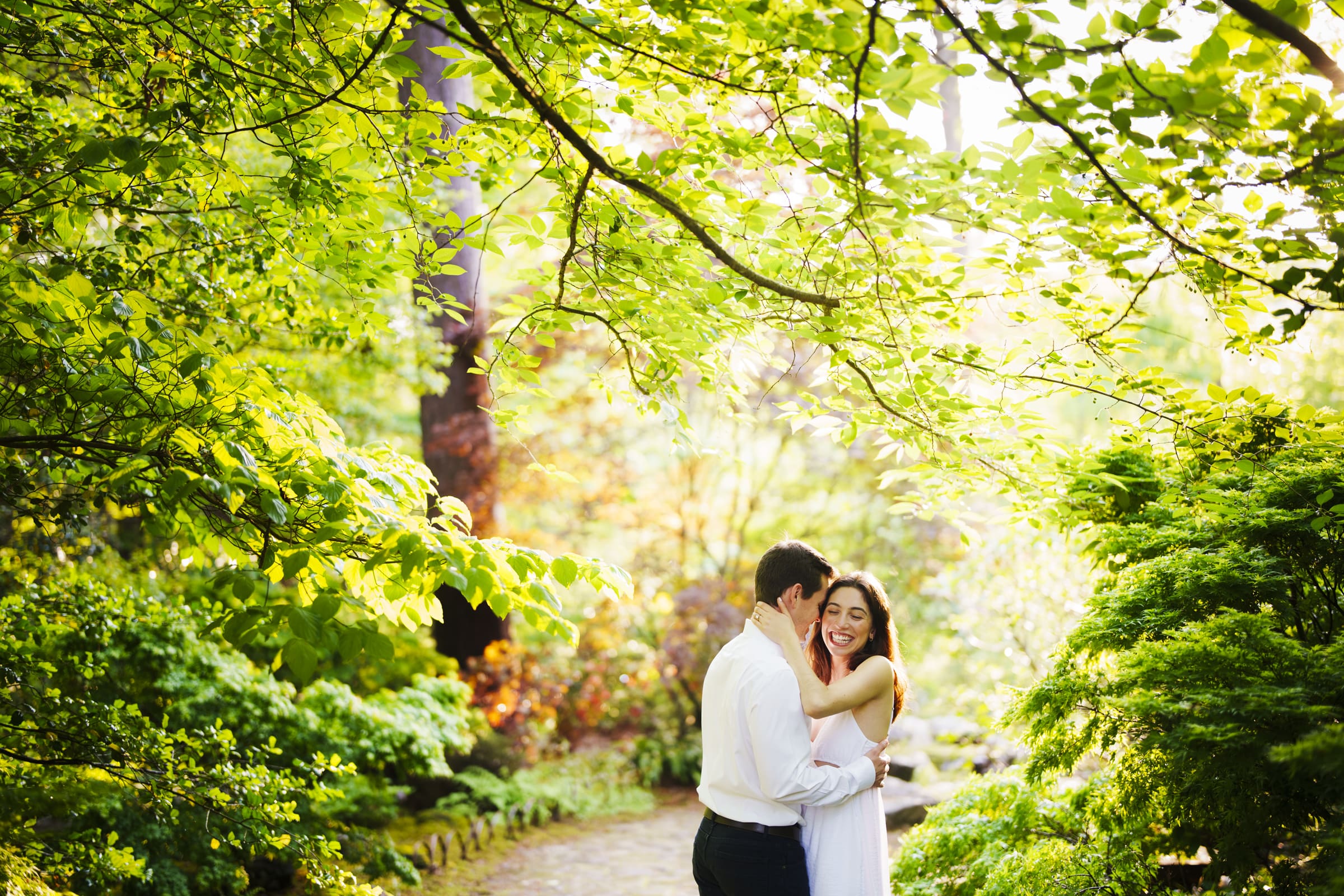 Gabby and Neil share a tender embrace under the lush green foliage of Duke Gardens. He wears a crisp white shirt, she a flowing white dress, as sunlight filters through the trees, casting a serene glow over their engagement session.