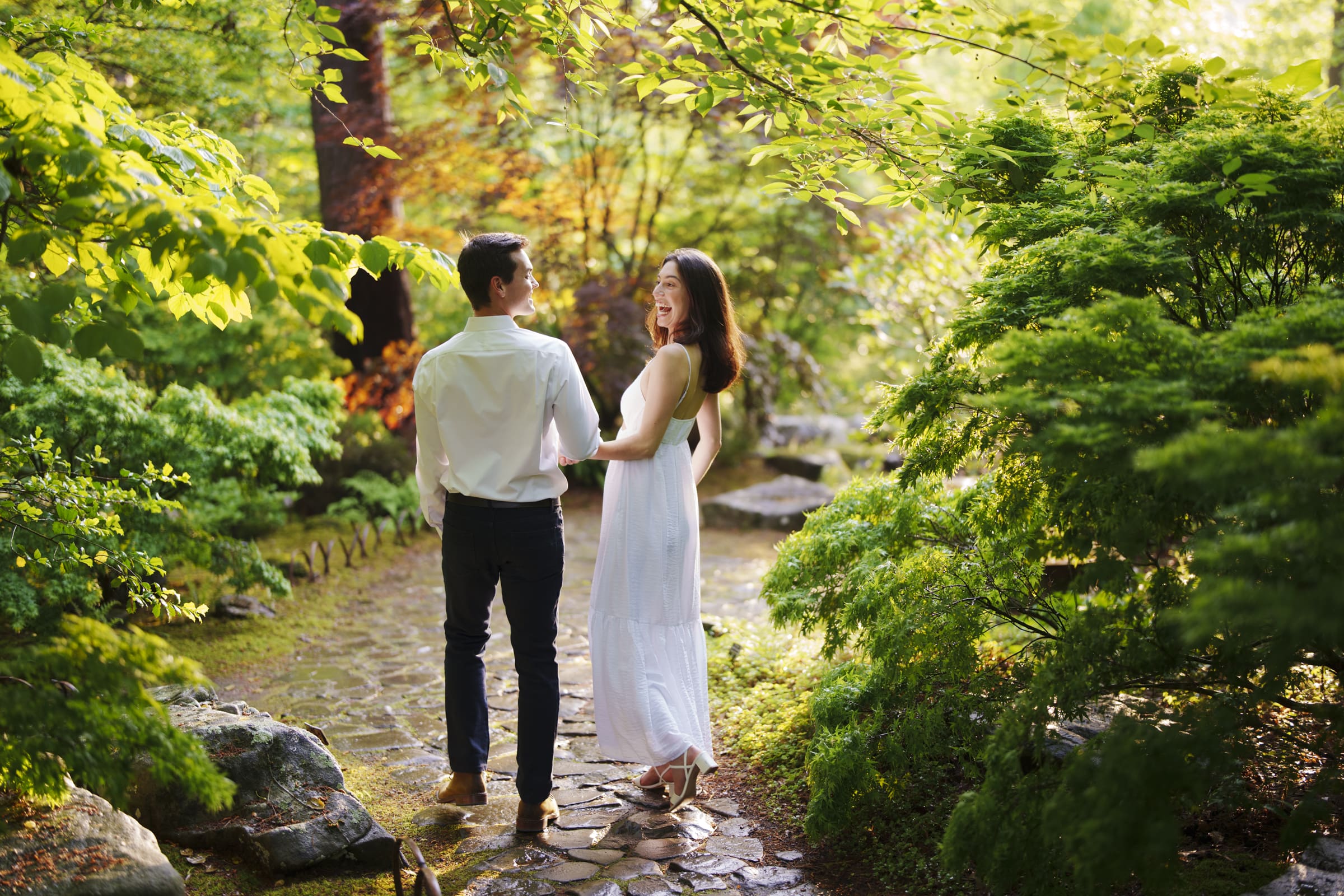 A couple strolls hand in hand along a stone path in a lush, sunlit garden during Gabby & Neil's Duke Gardens engagement session. The woman wears a white dress, and the man is in a white shirt and dark pants. They both appear happy amidst vibrant greenery.
