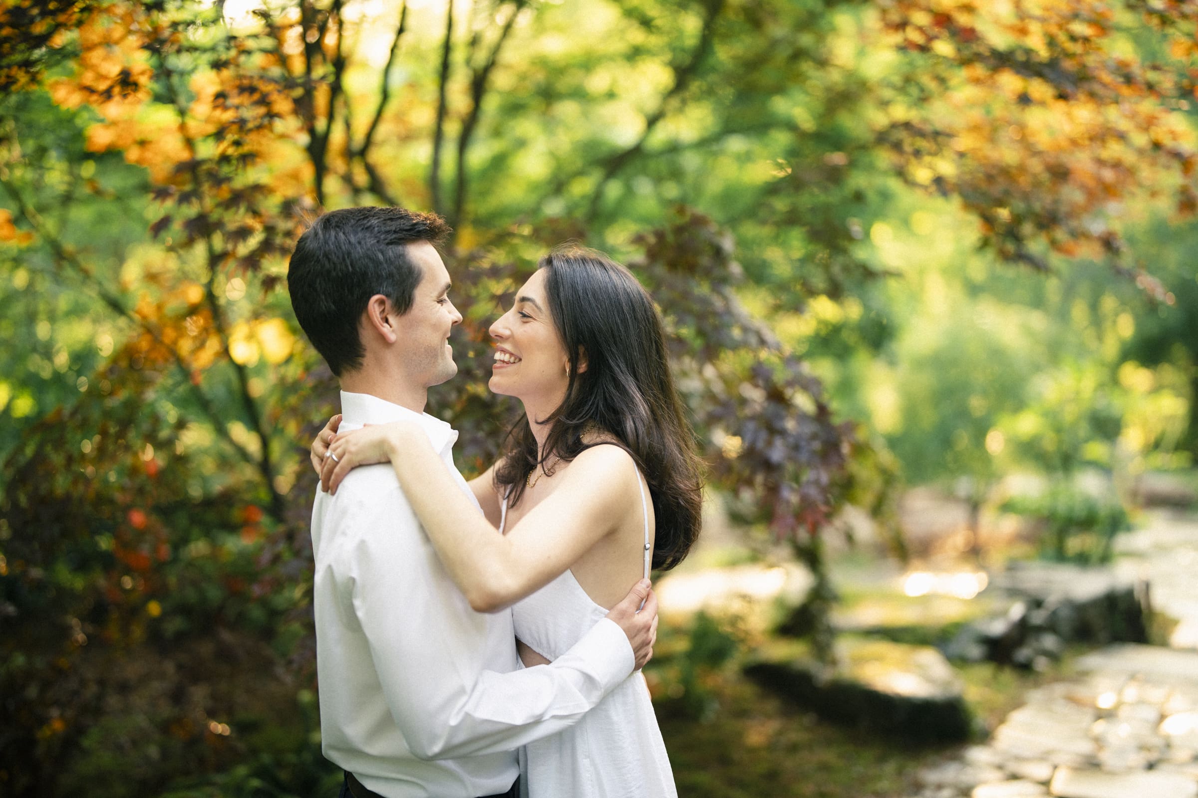 Gabby and Neil embrace and smile at each other in a lush, sunlit garden with vibrant green and orange foliage. Their Duke Gardens engagement session is serene and joyful, capturing a moment of connection amidst nature.