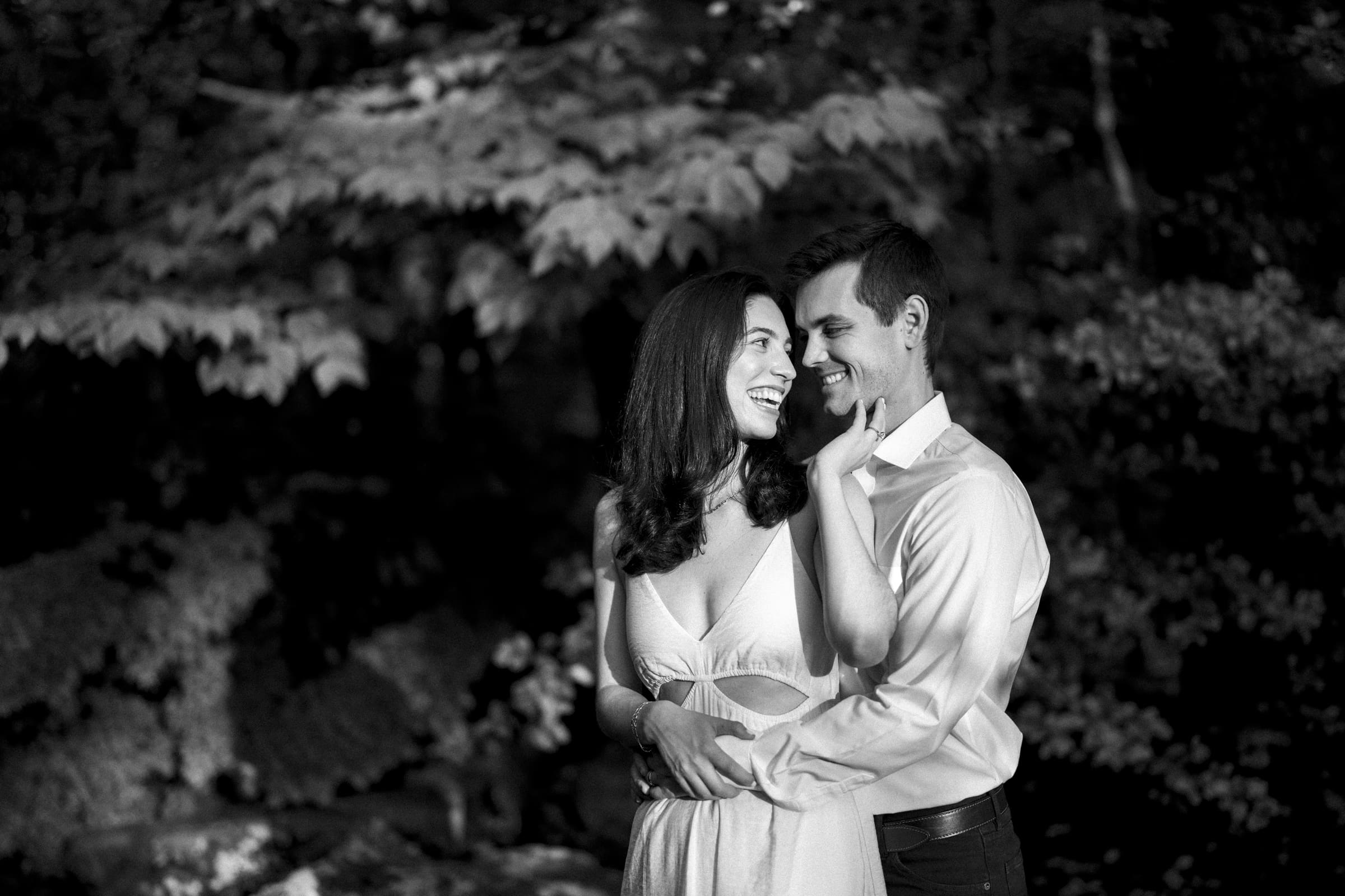 In this enchanting Duke Gardens engagement session, Gabby and Neil embrace and smile under the dappled sunlight. She, in a light dress, gently touches his face while he, dressed in a button-up shirt, wraps an arm around her waist amidst the serene beauty of the woods.