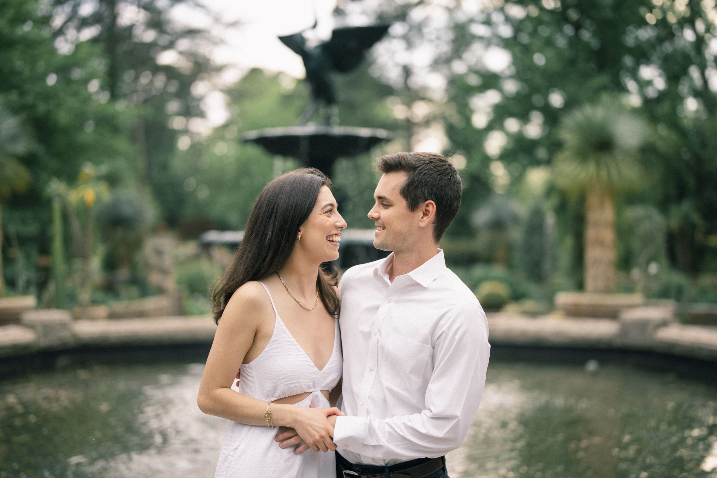 Gabby and Neil share a loving smile during their engagement session at Duke Gardens. Both dressed in white, they stand by a fountain surrounded by lush greenery, her long dark hair flowing gracefully, his short dark hair complementing the tranquil water scene in the background.